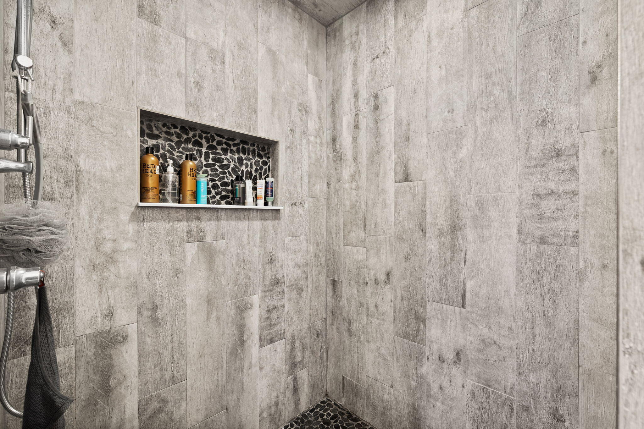 Tiled shower with a pebble-mosaic niche and floor, toiletries, and a handheld showerhead.