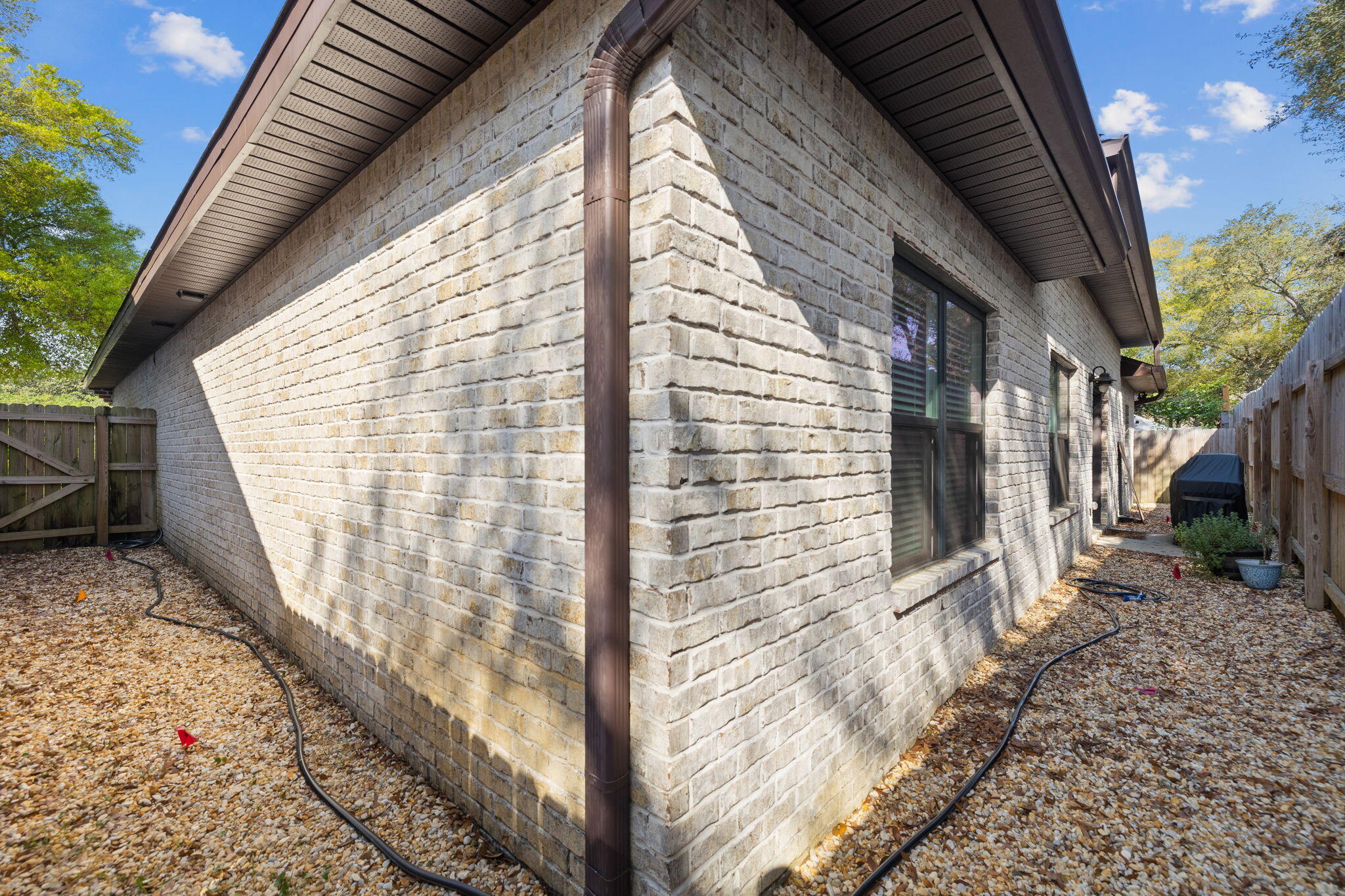 Light brick house side with dark-framed windows and a gravel path between wooden fences.