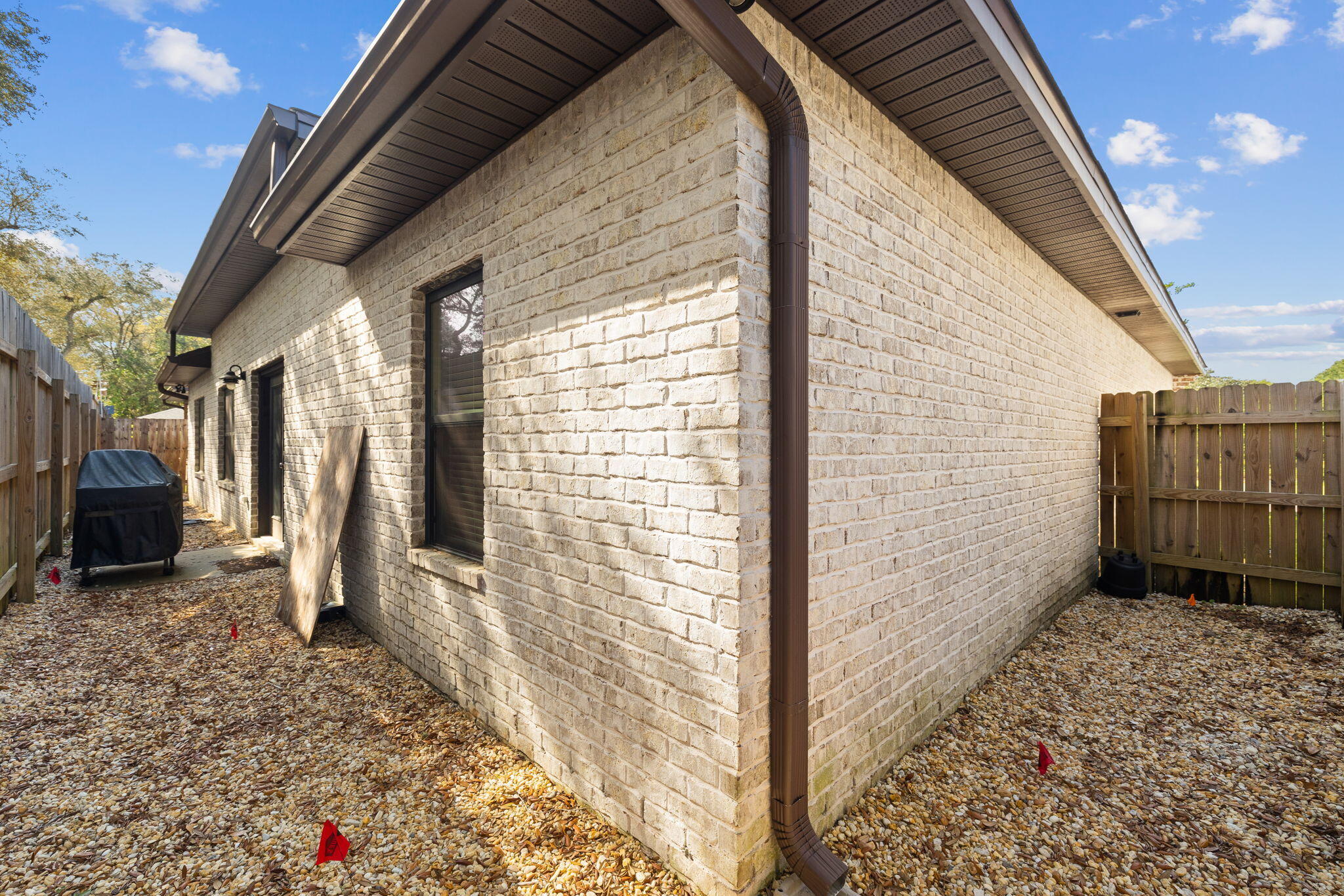 Light brick side of the home with dark gutters and a fenced gravel yard.