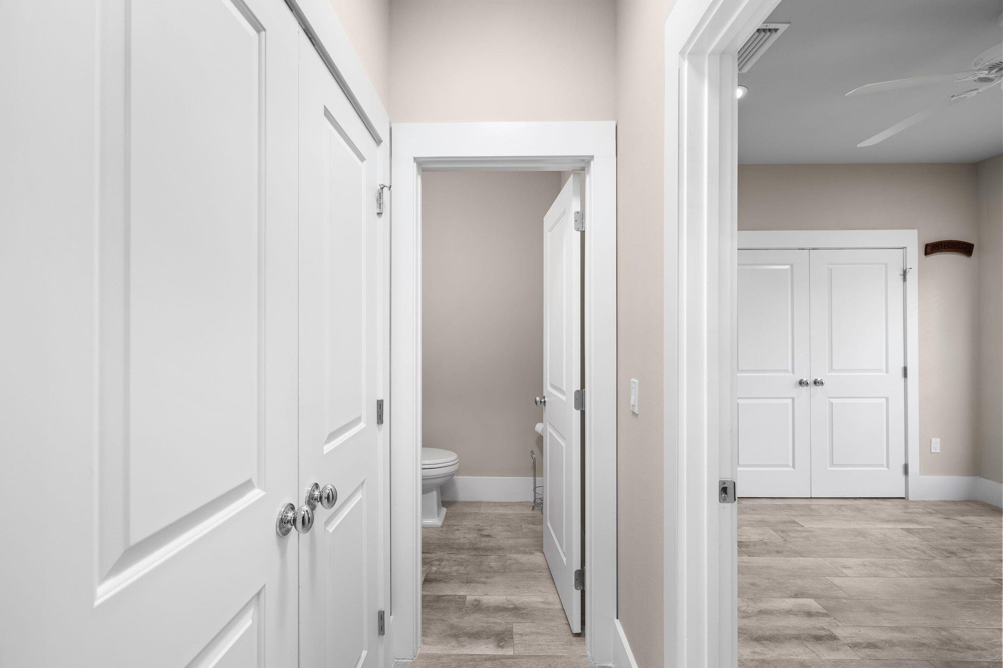 Bathroom with a toilet seen through open doors beside an empty tiled room with a ceiling fan.