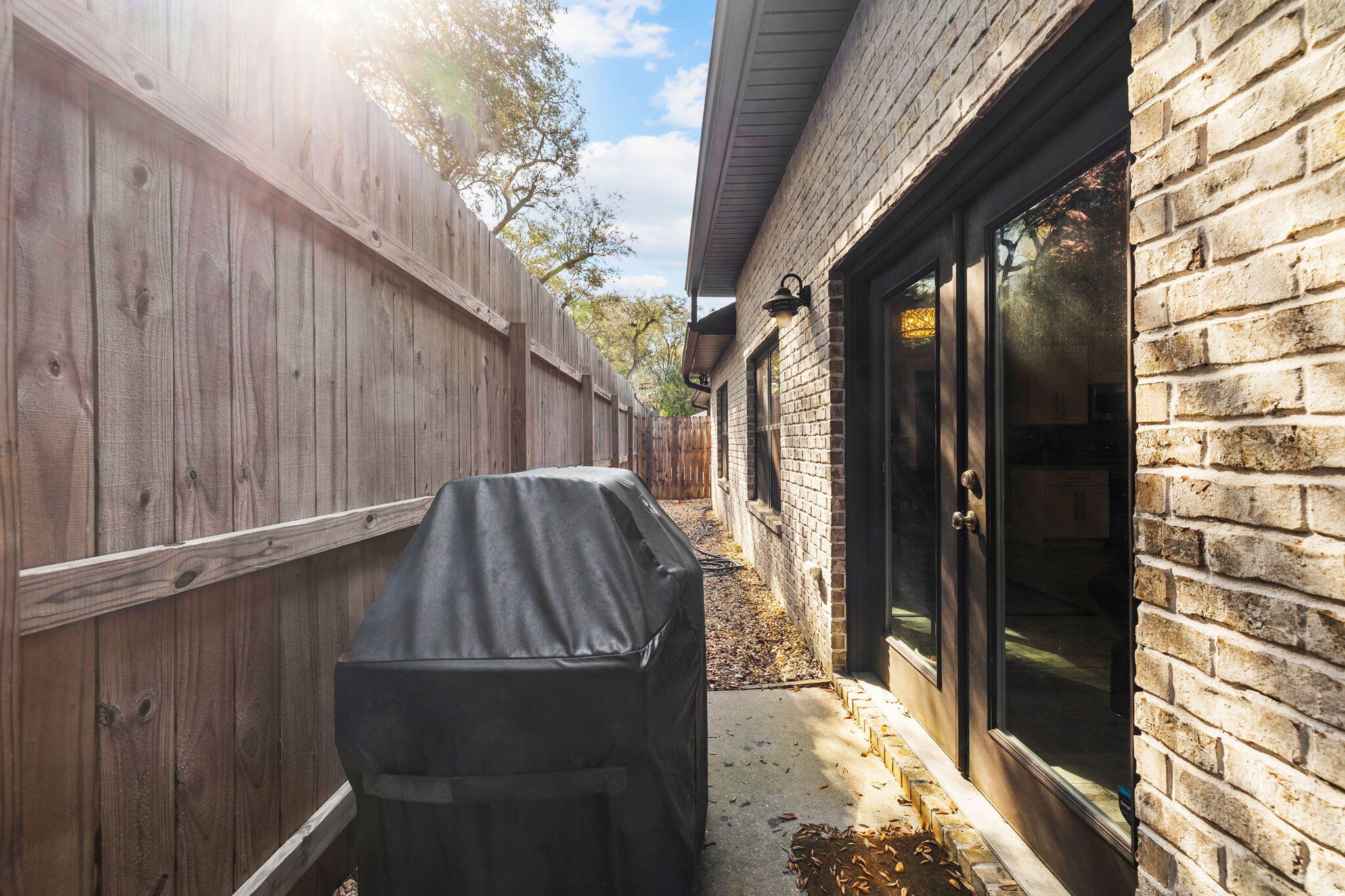 Side patio with a covered grill, wooden privacy fence, and black-framed glass doors.
