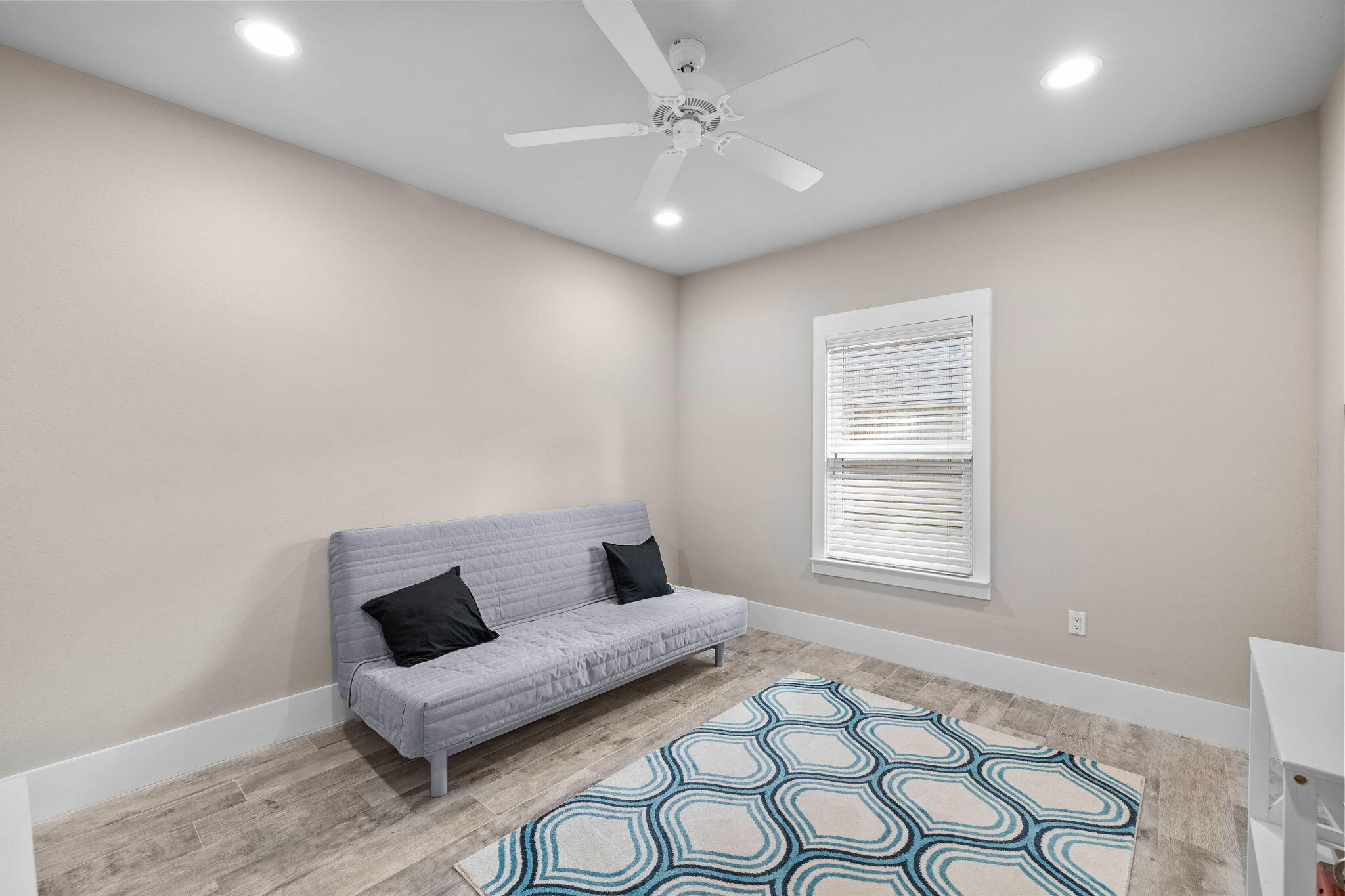 Room with gray futon, blue patterned rug, wood-look tile floor, and window with blinds.