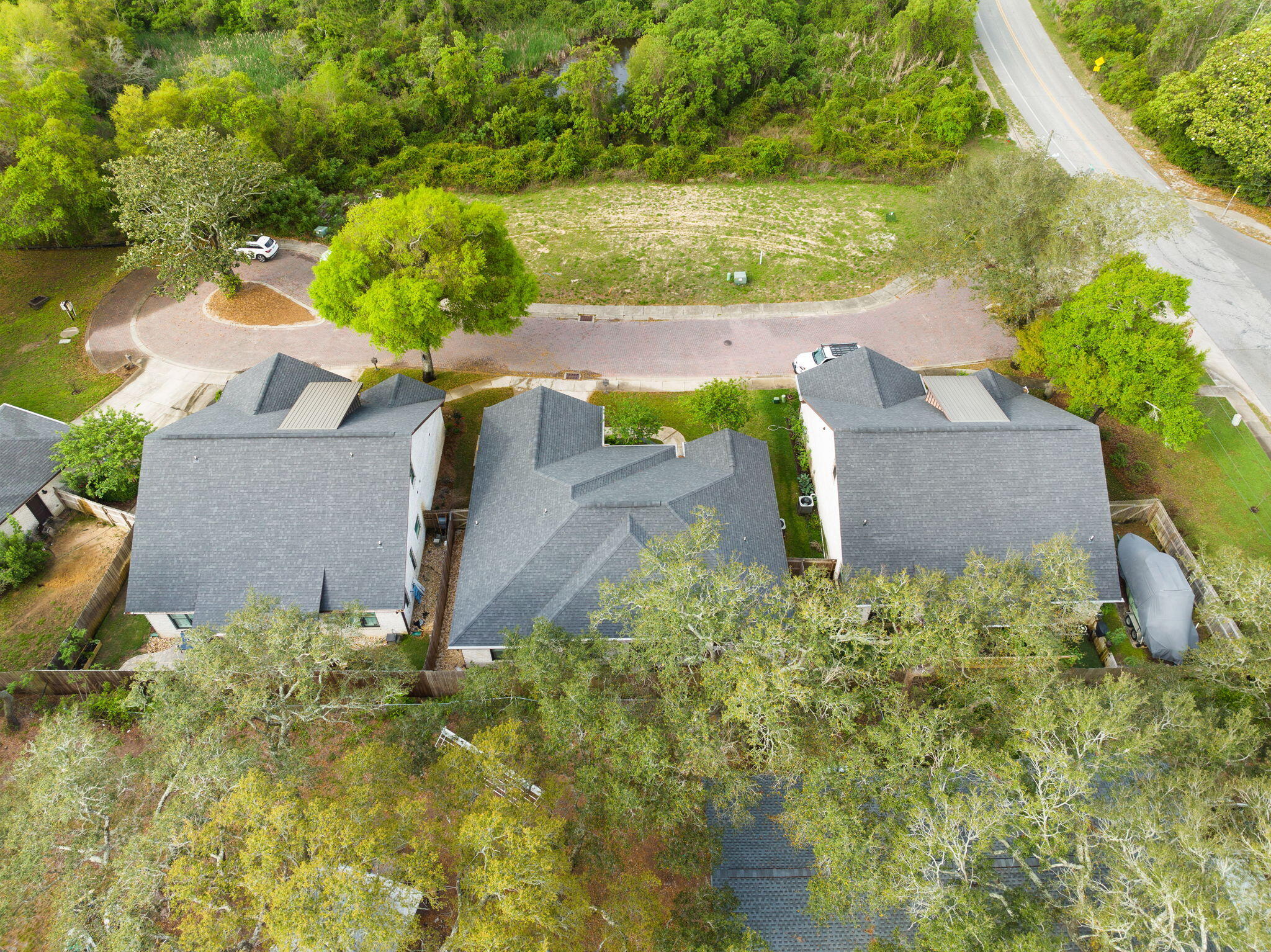 Aerial view of homes with gray roofs, fenced yards, a brick-paved street, and surrounding trees.