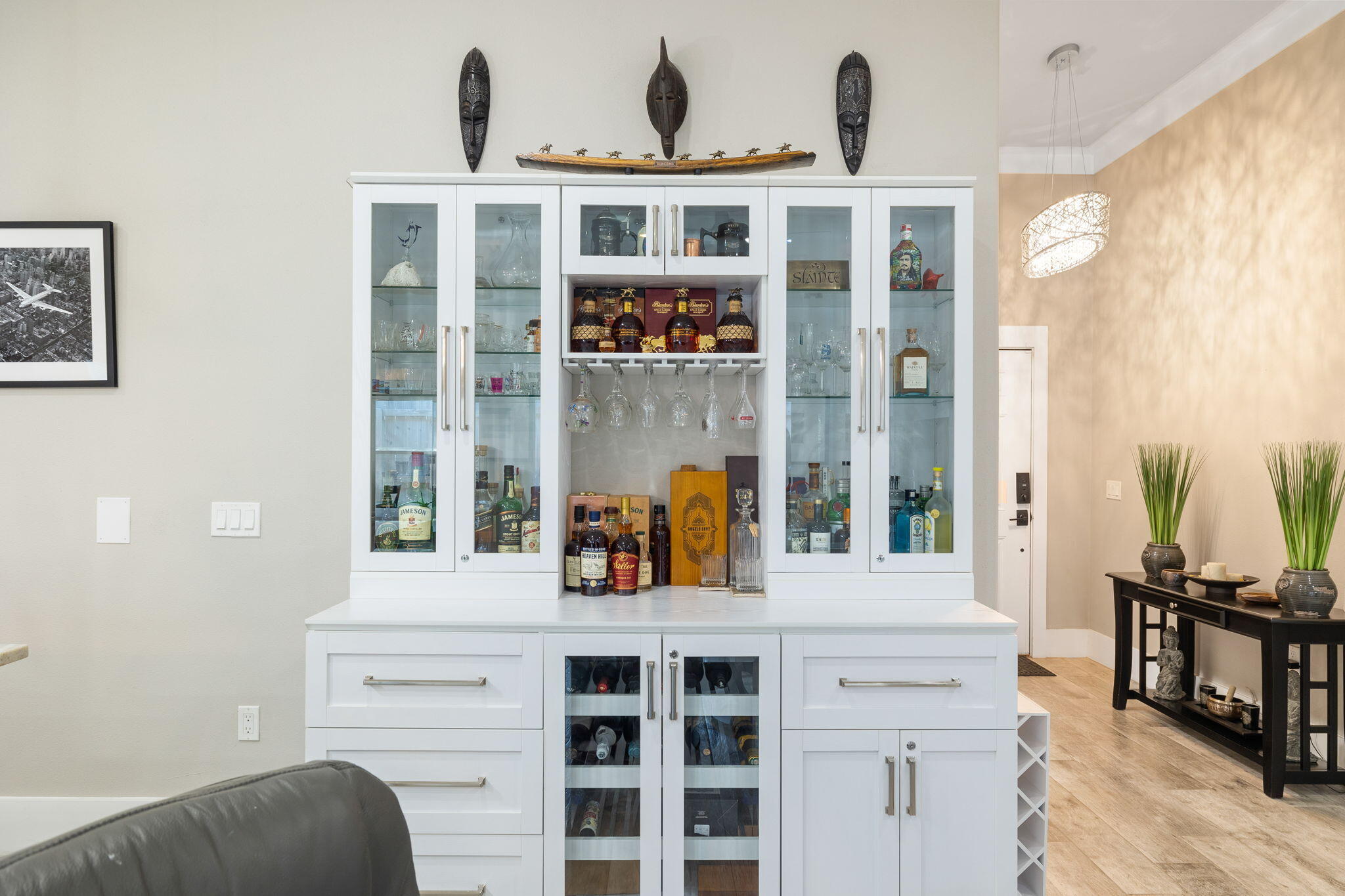 White built-in bar cabinet with glass-front cupboards, liquor bottles, and a wine fridge beside a pendant-lit hallway.