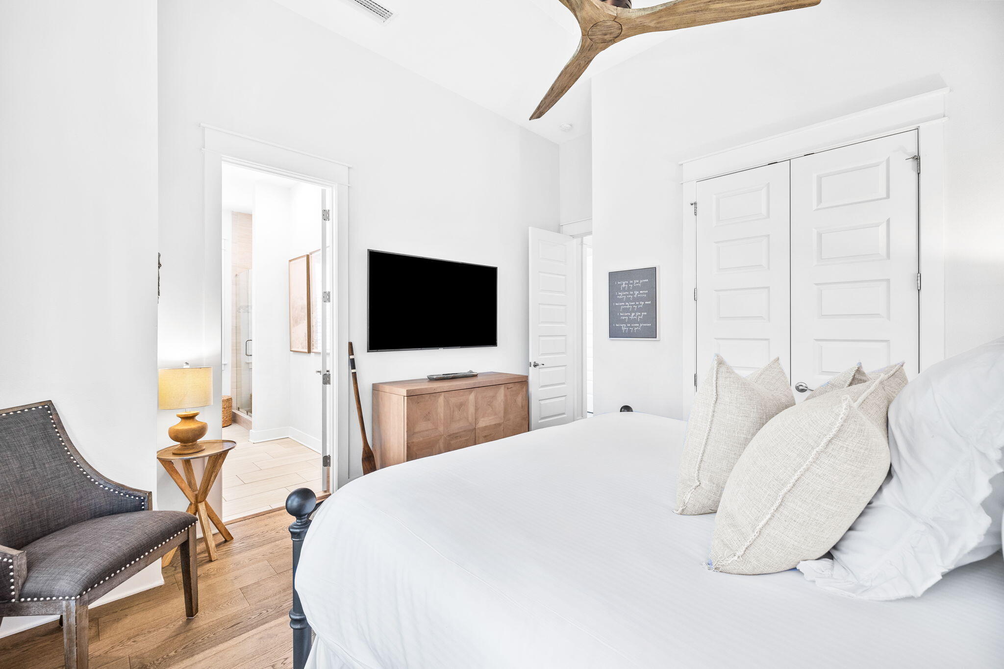 Bedroom with a large white bed, wood floors, a dark chair, and a television above a wooden dresser.