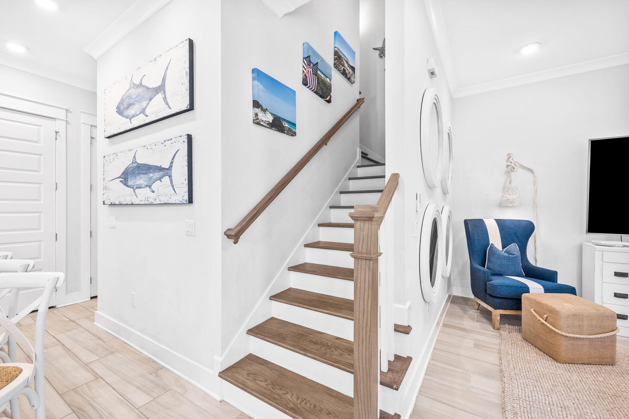 Interior staircase with wood treads, coastal artwork, and a blue armchair in a bright sitting area.