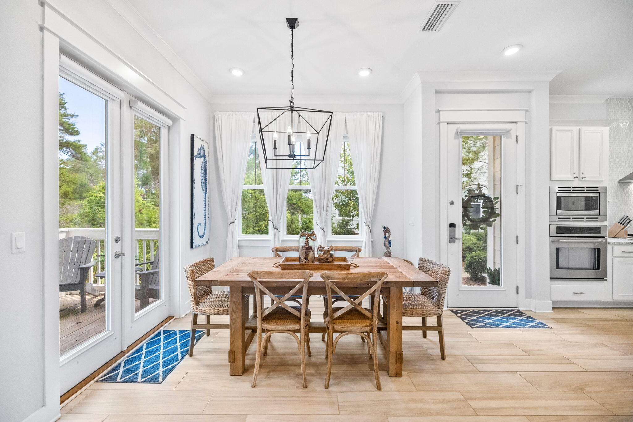 Dining room with a rustic wood table, woven chairs, and large windows overlooking trees.