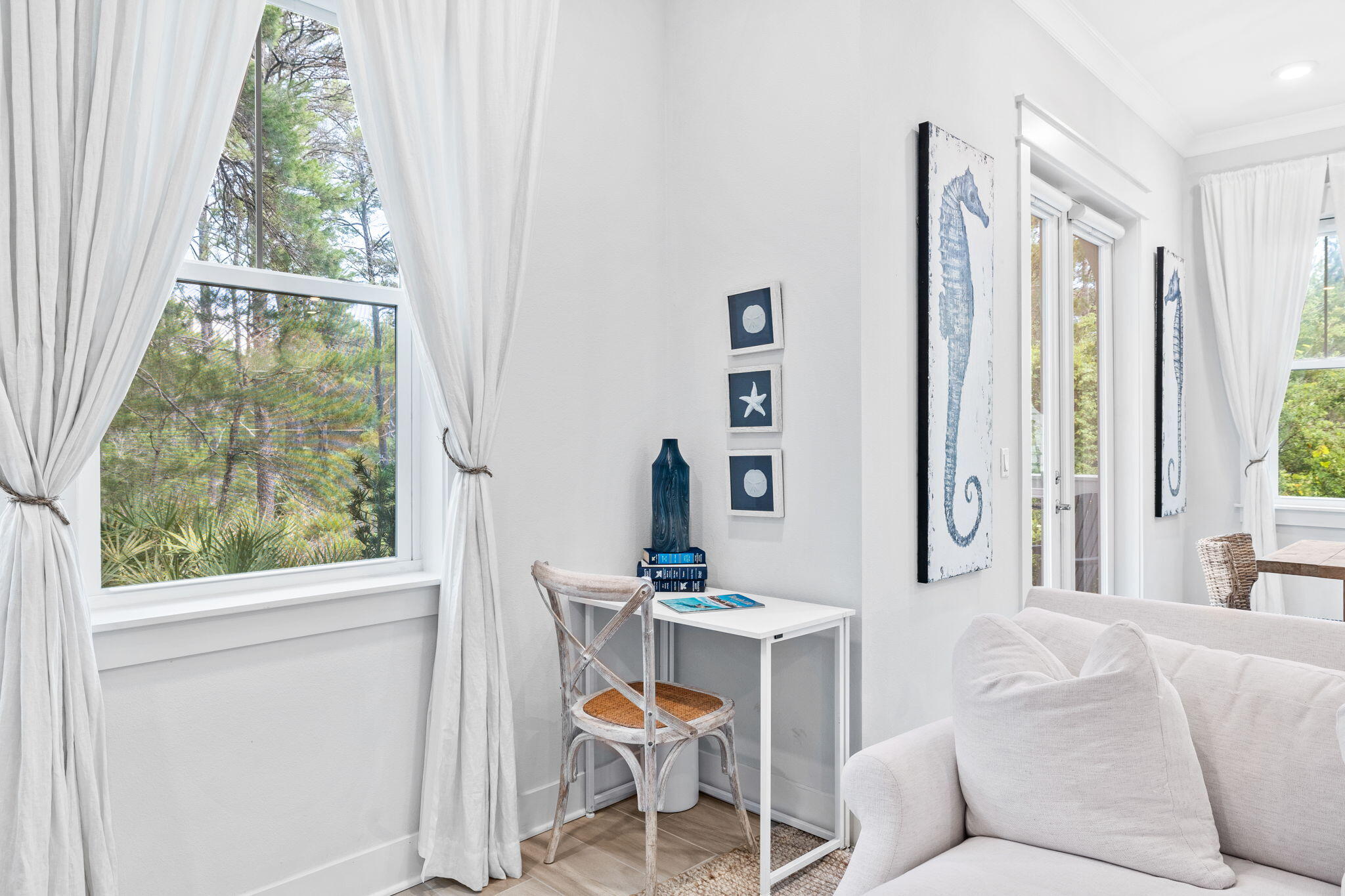 Bright living area with large curtained windows, a small desk, and blue seahorse artwork.