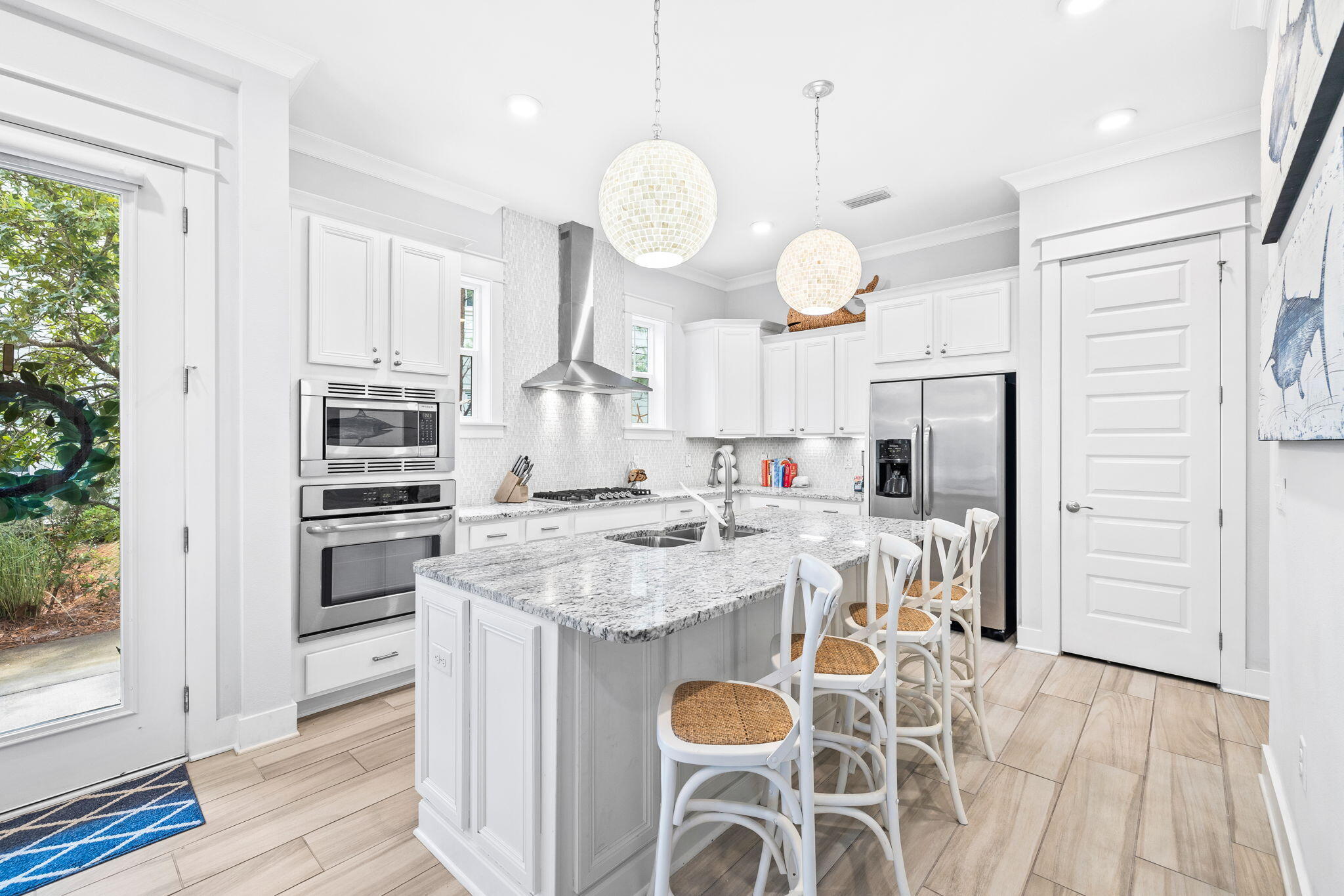 Bright white kitchen with a large granite island, woven-seat stools, and stainless steel appliances.