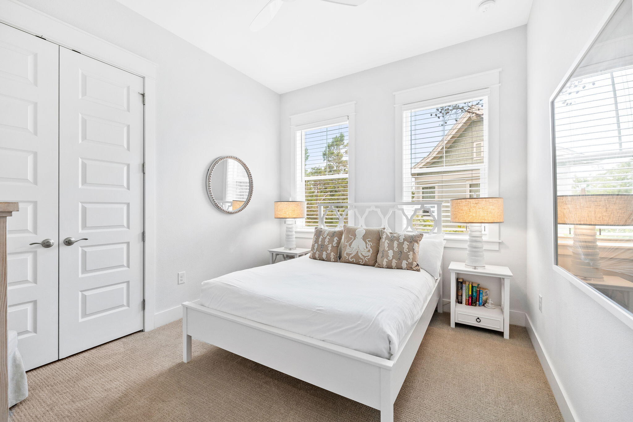 Bright bedroom with a white bed, patterned pillows, and windows overlooking a neighboring house.