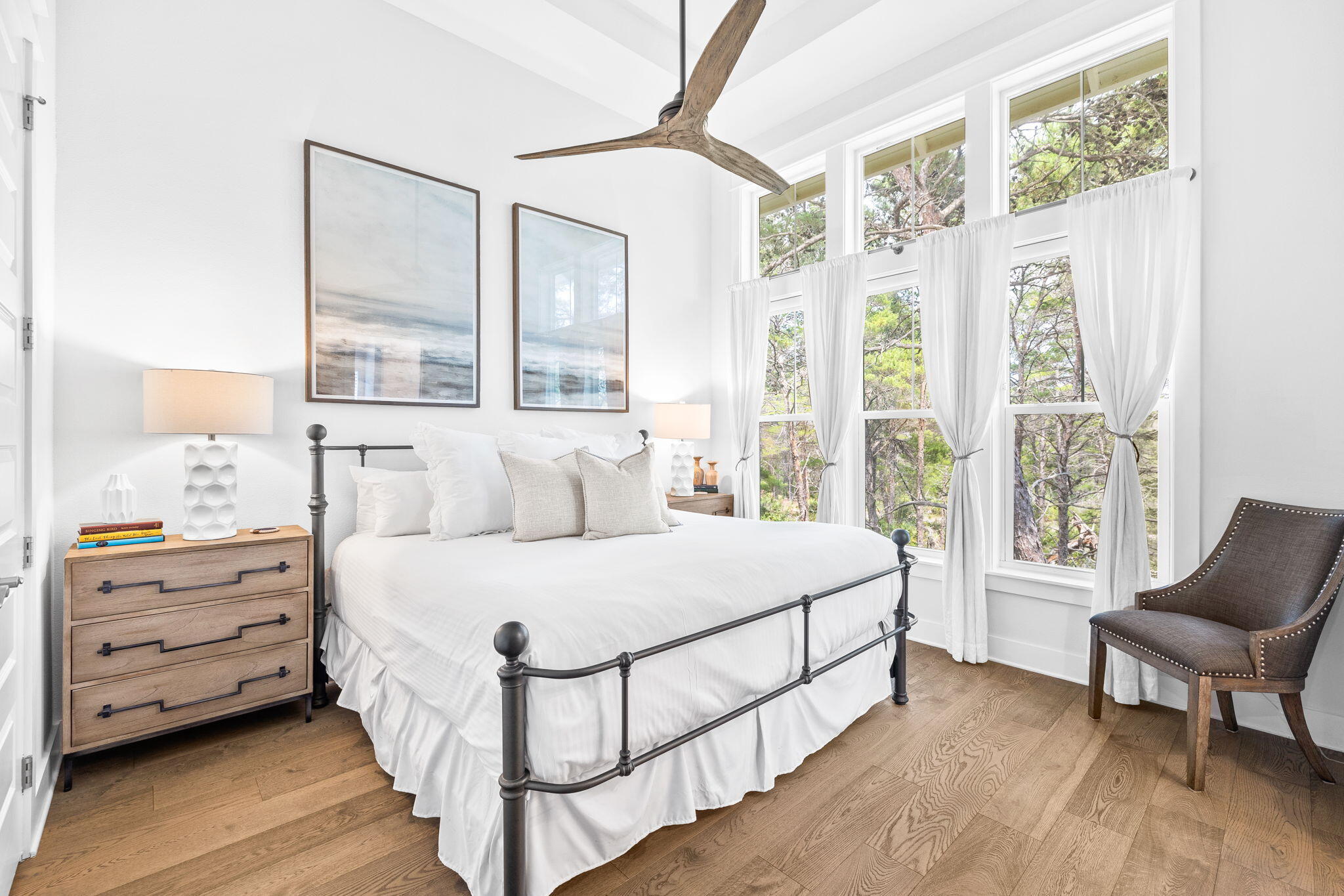 Bright bedroom with a white bed, wood floors, and tall curtained windows overlooking trees.
