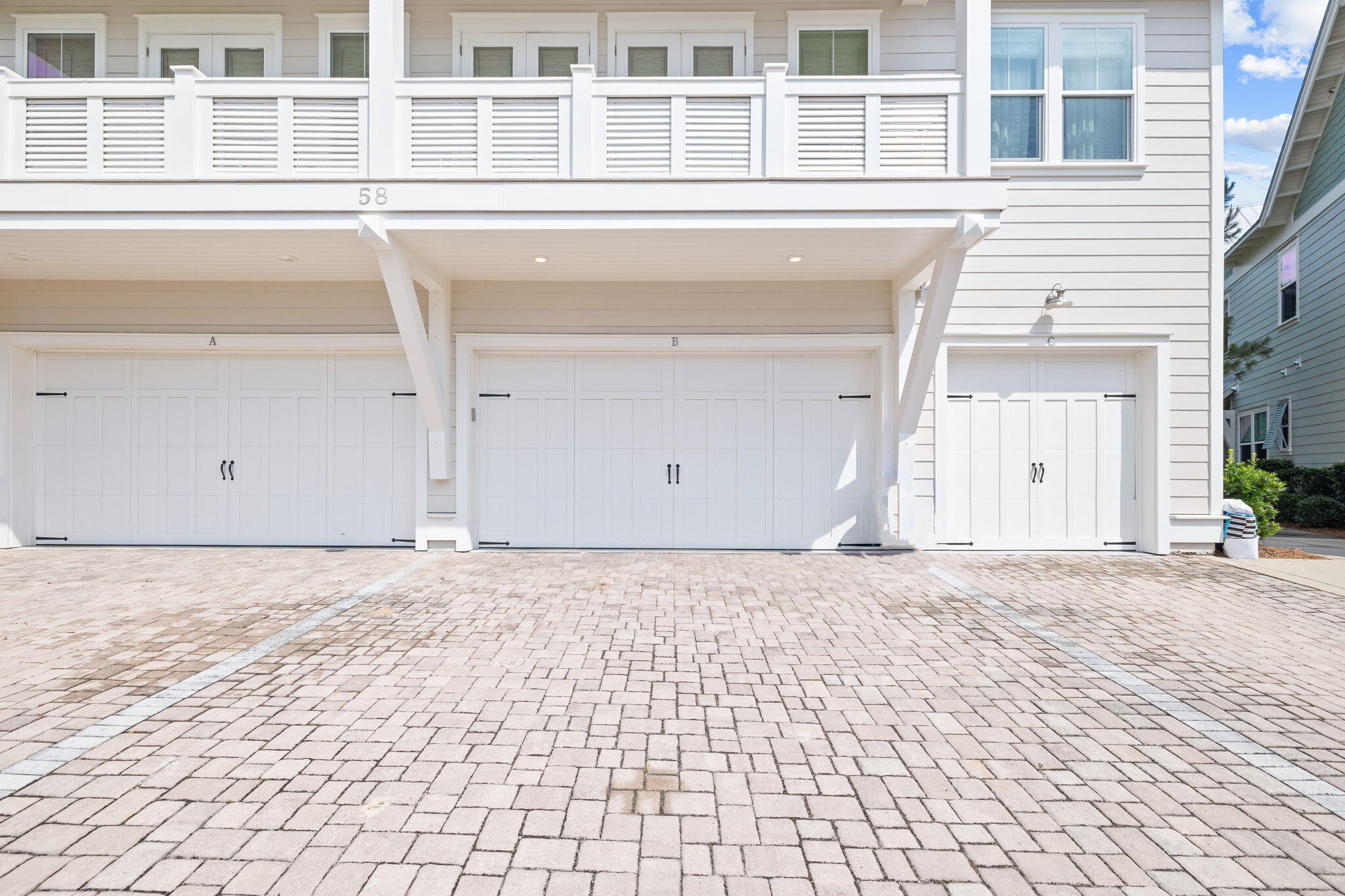 White multi-unit building with three garage doors, an upper balcony, and a wide paver driveway.
