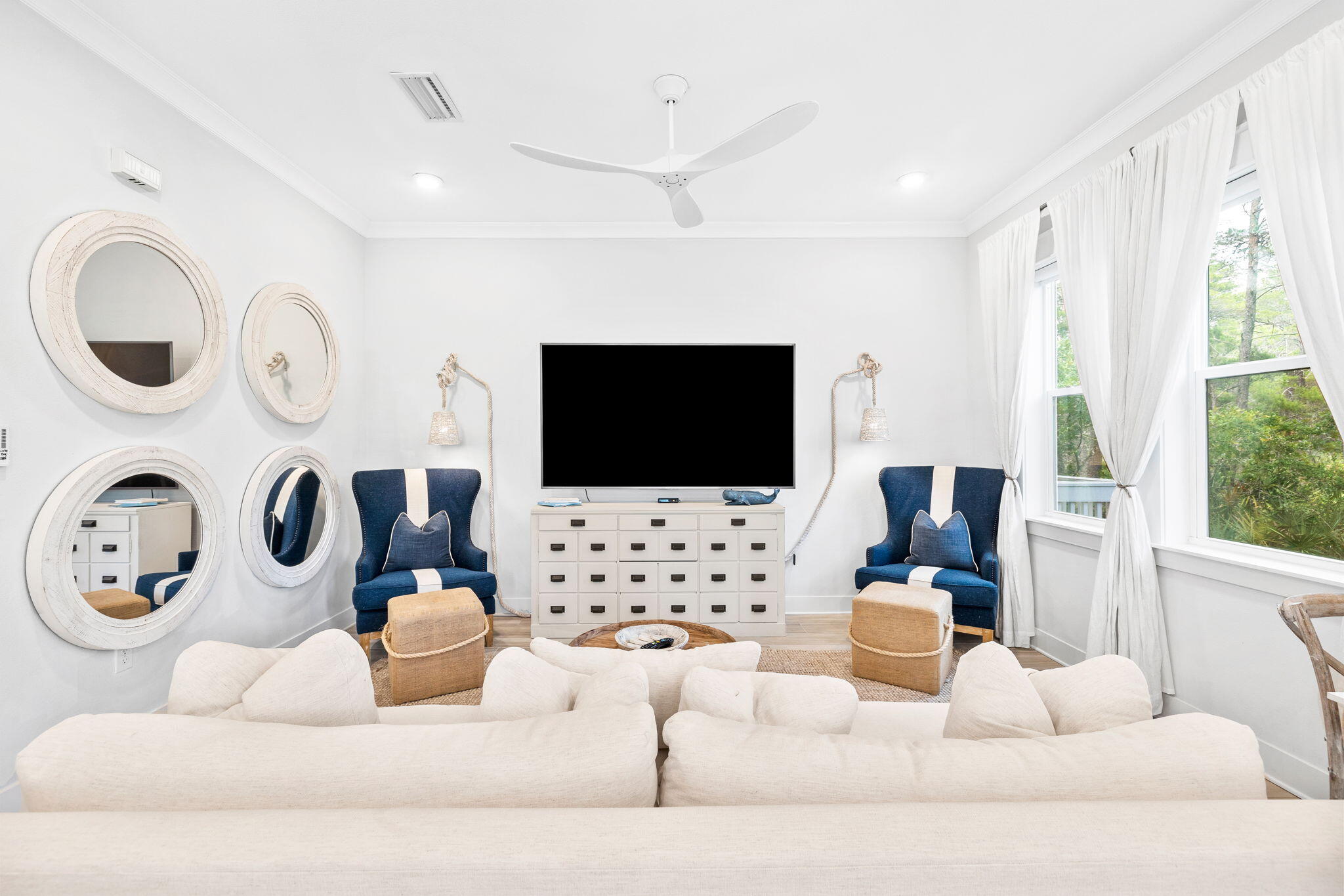 Bright living room with white sofas, navy armchairs, round mirrors, and tall windows overlooking greenery.
