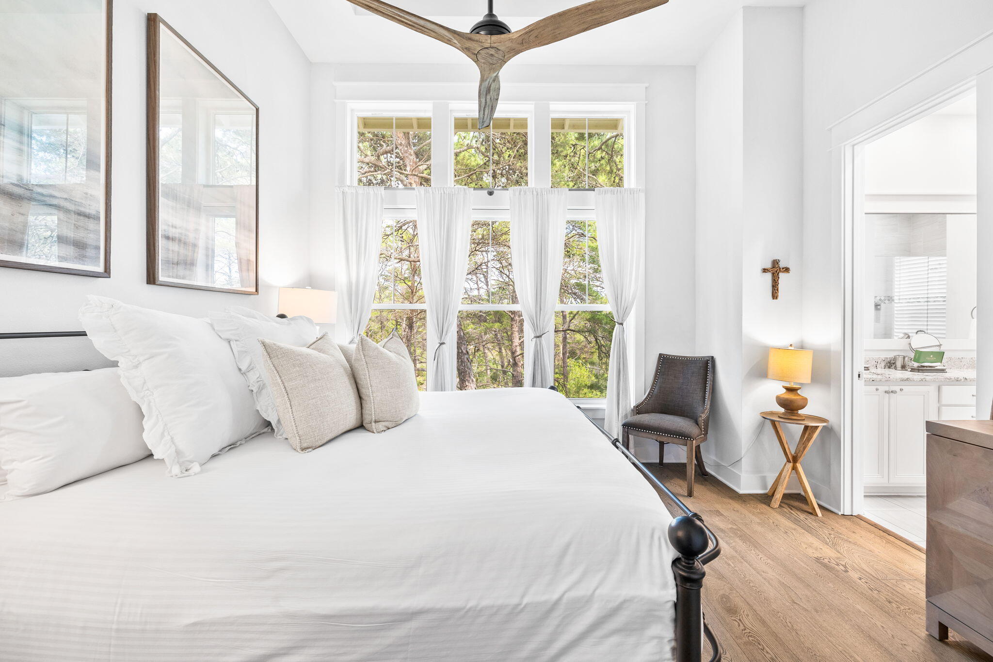 Bright bedroom with a large white bed, tall curtained windows, and a wood ceiling fan.