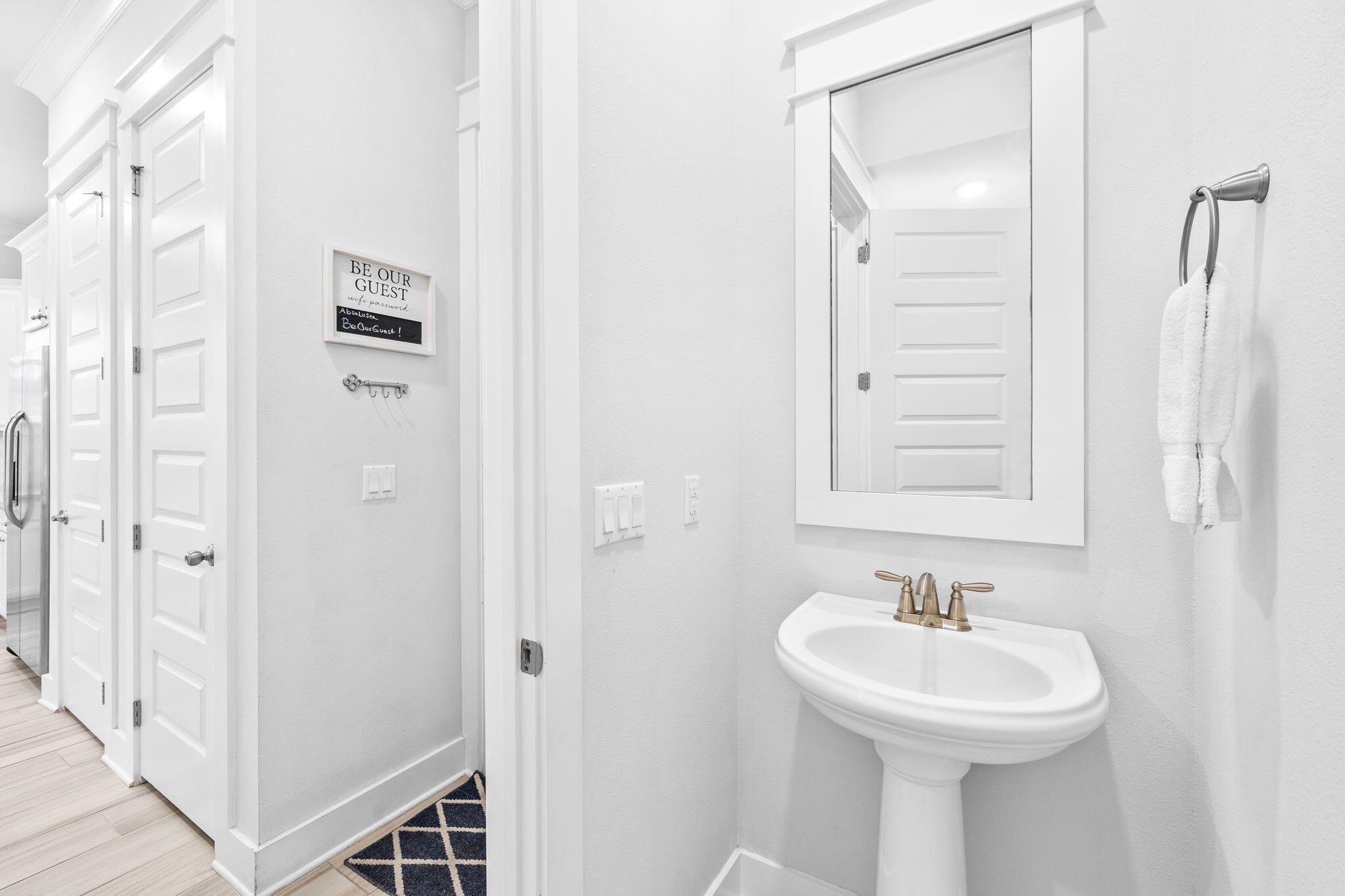 Bright white bathroom with a pedestal sink, brass faucet, framed mirror, and guest welcome sign.