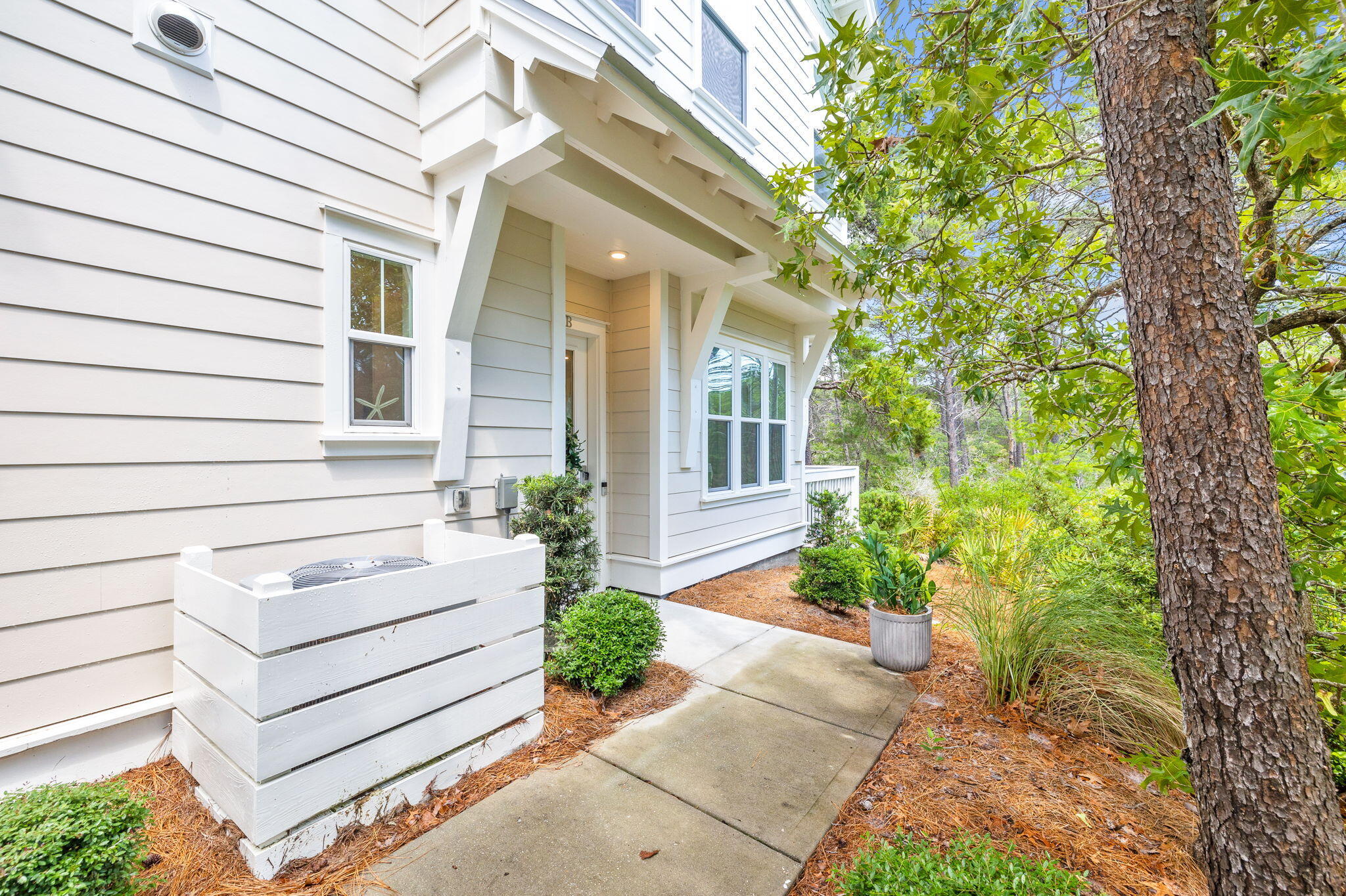 Home entrance with a covered porch, concrete walkway, and landscaped shrubs beneath a large tree.
