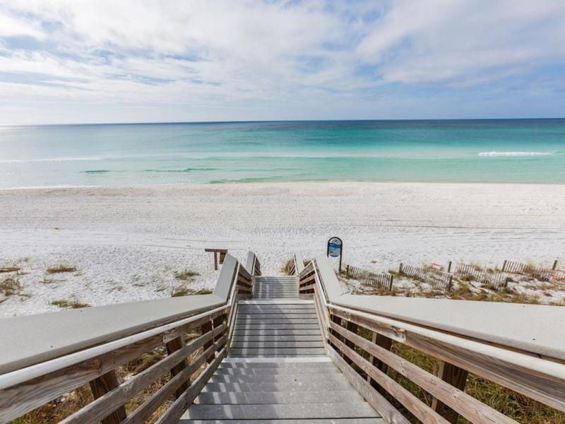 Boardwalk across white sand leads to turquoise water beneath a cloudy sky.