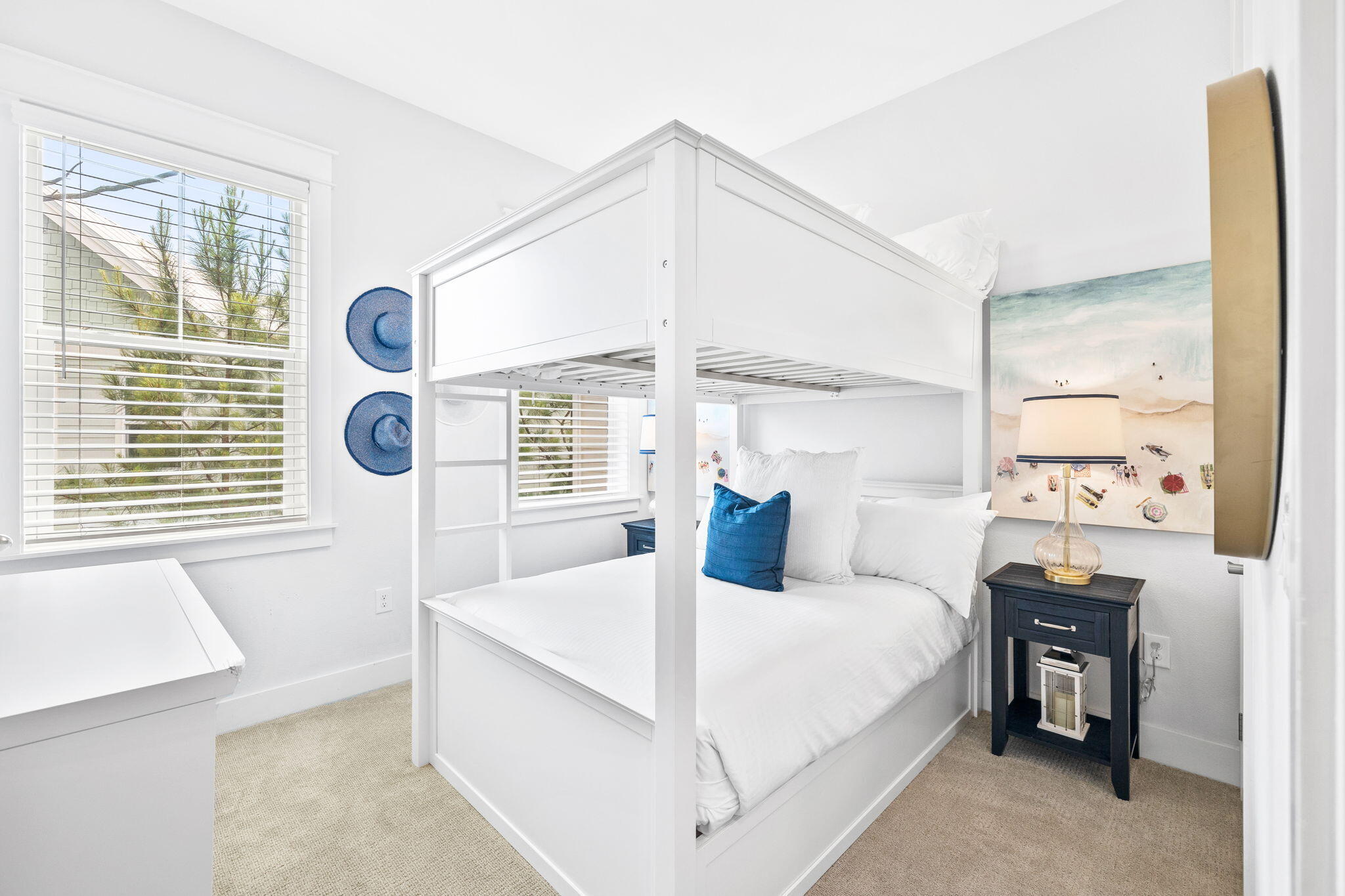 Bright bedroom with a white bunk bed, blue accent pillows, coastal artwork, and large windows with blinds.