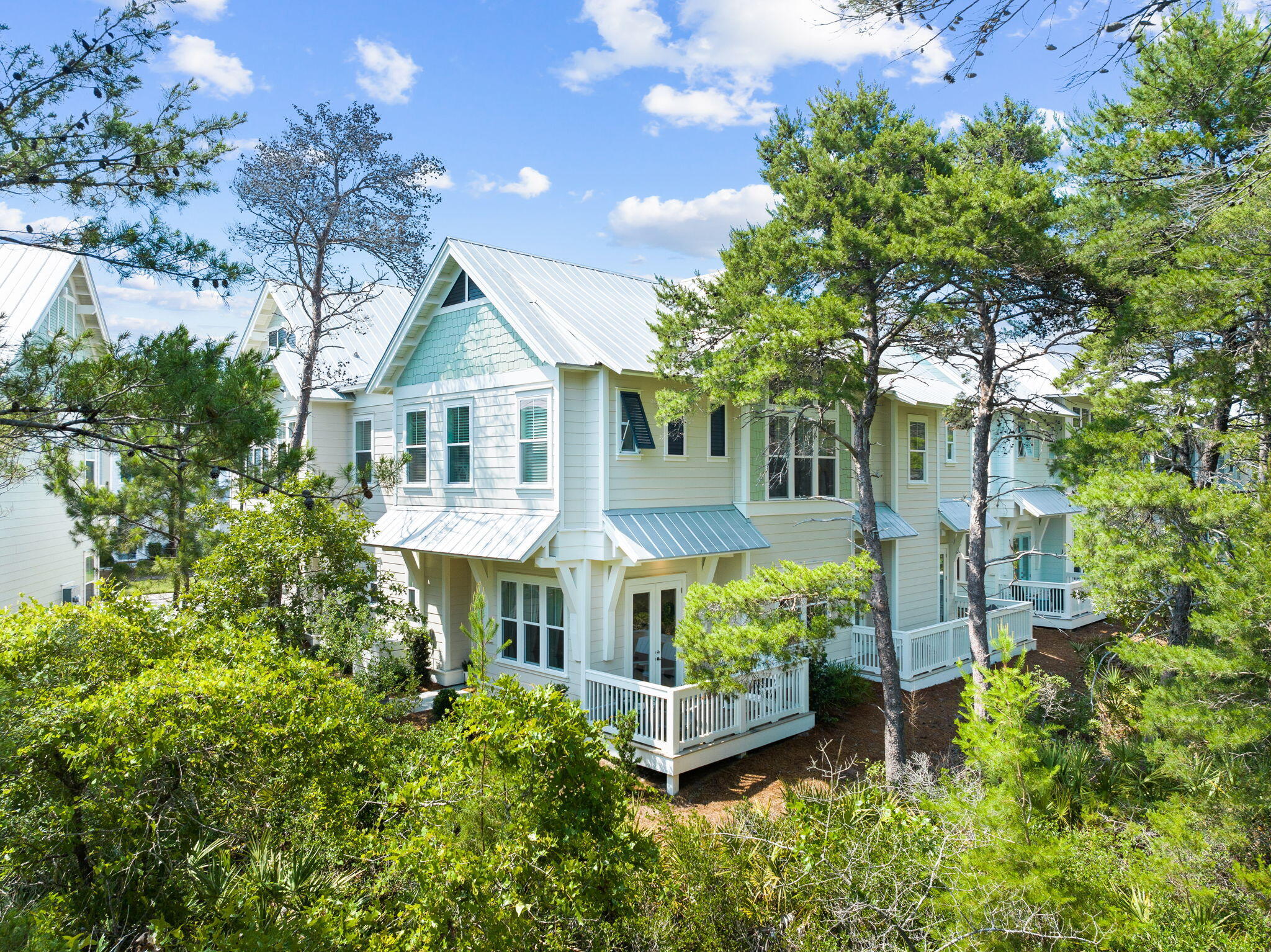 Coastal-style home with white metal gabled roofs and porches among pine trees and dense greenery.