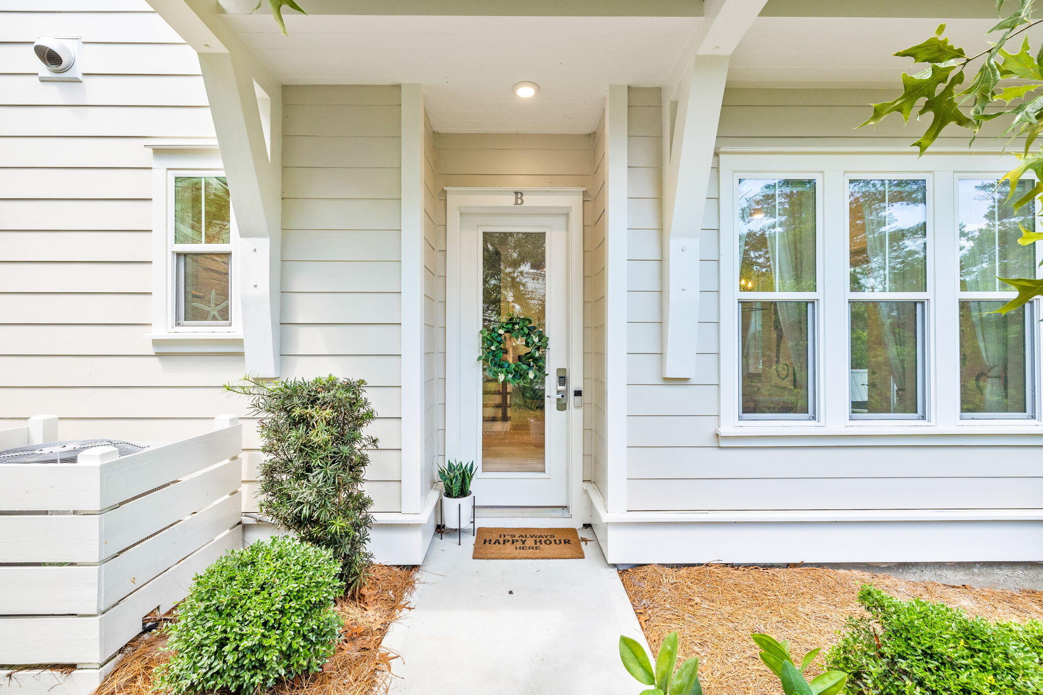 Covered entrance to Unit B with white siding, a glass-paneled door, and shrubs.