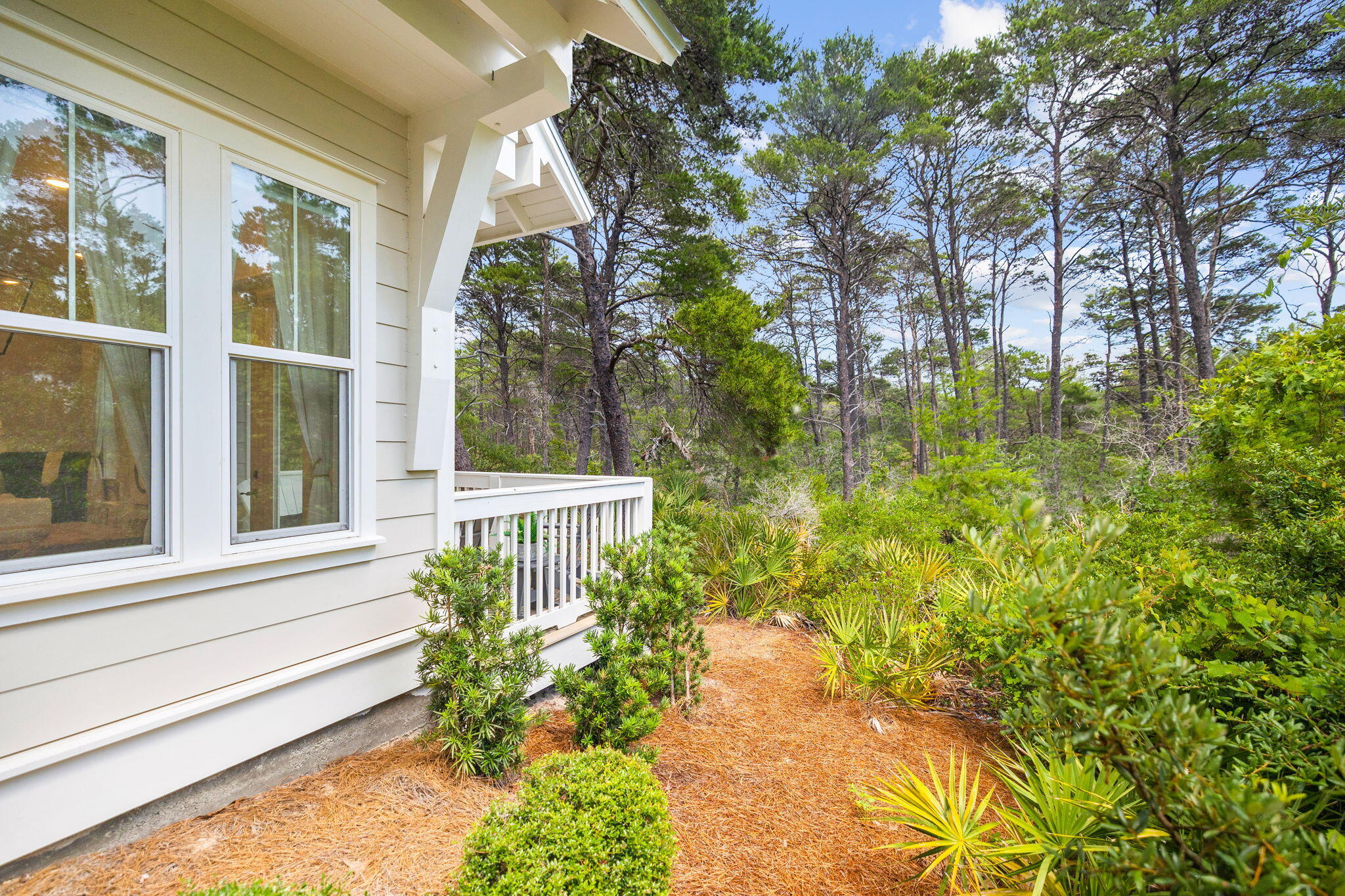 Light-colored home with a covered porch and large windows beside a wooded, landscaped yard.
