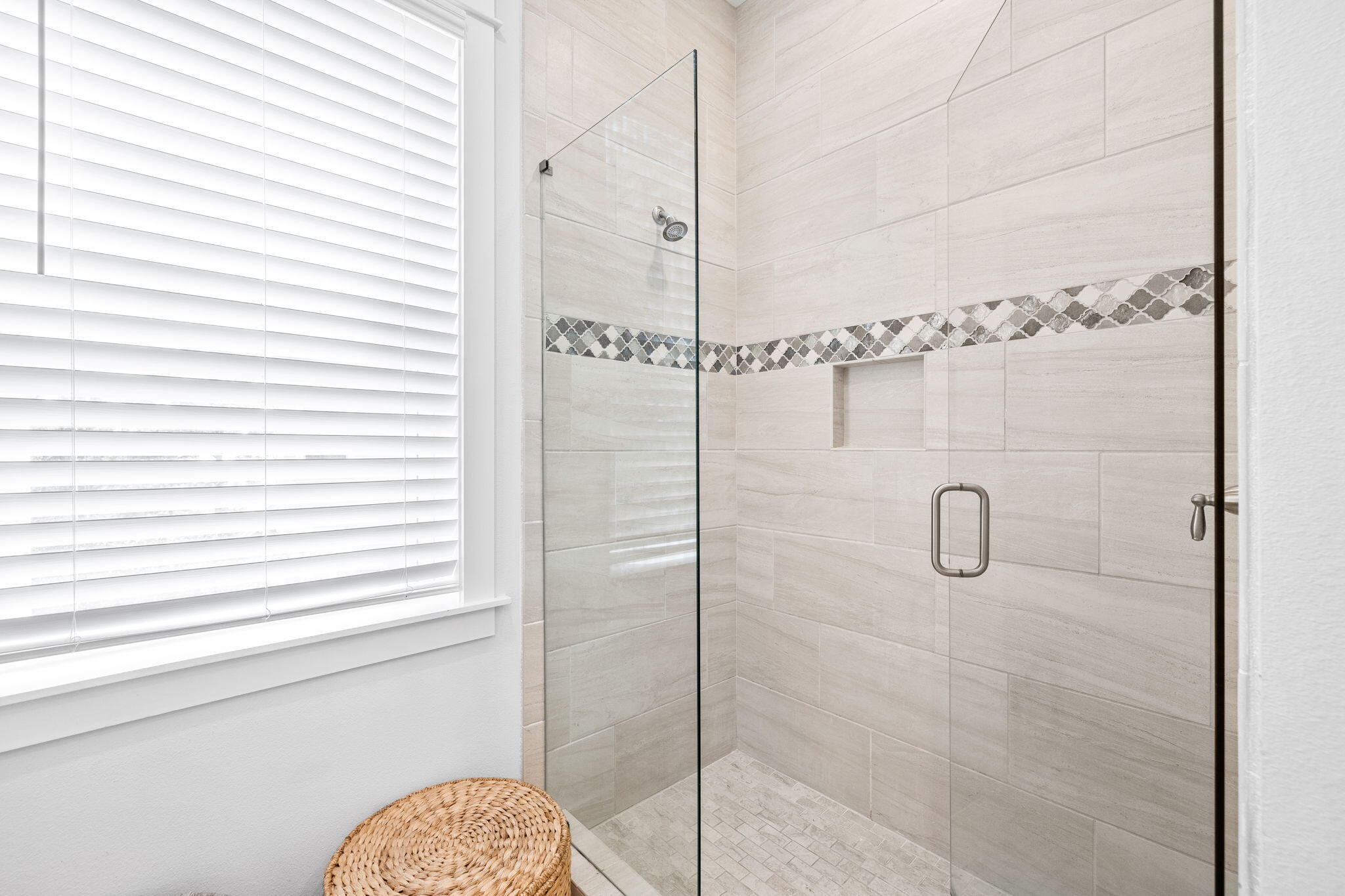 Walk-in shower with pale tile, patterned accent band, glass enclosure, and a window with white blinds.