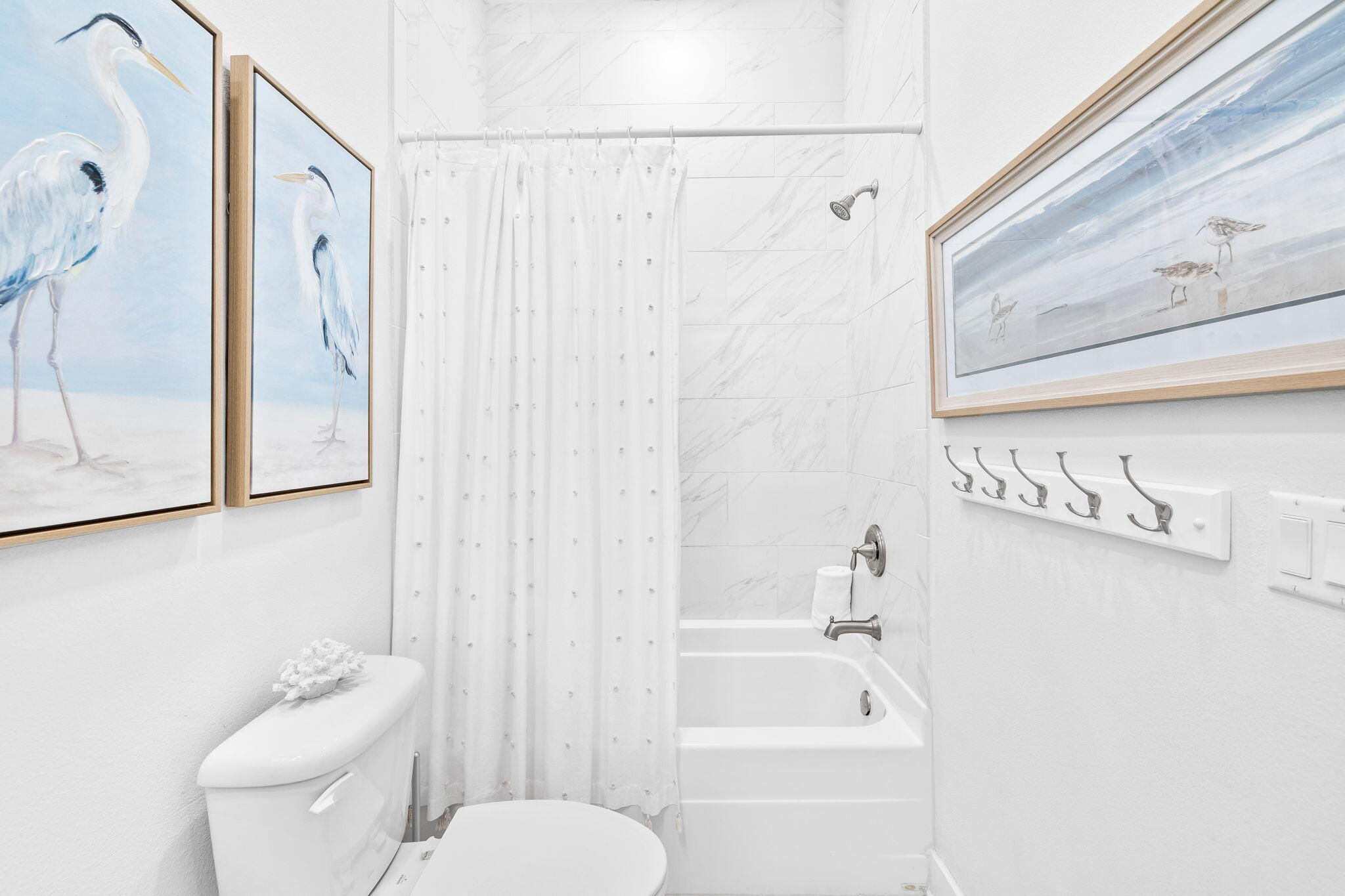 Bright bathroom with a white tub-shower, marble-look tile, and framed coastal bird artwork.