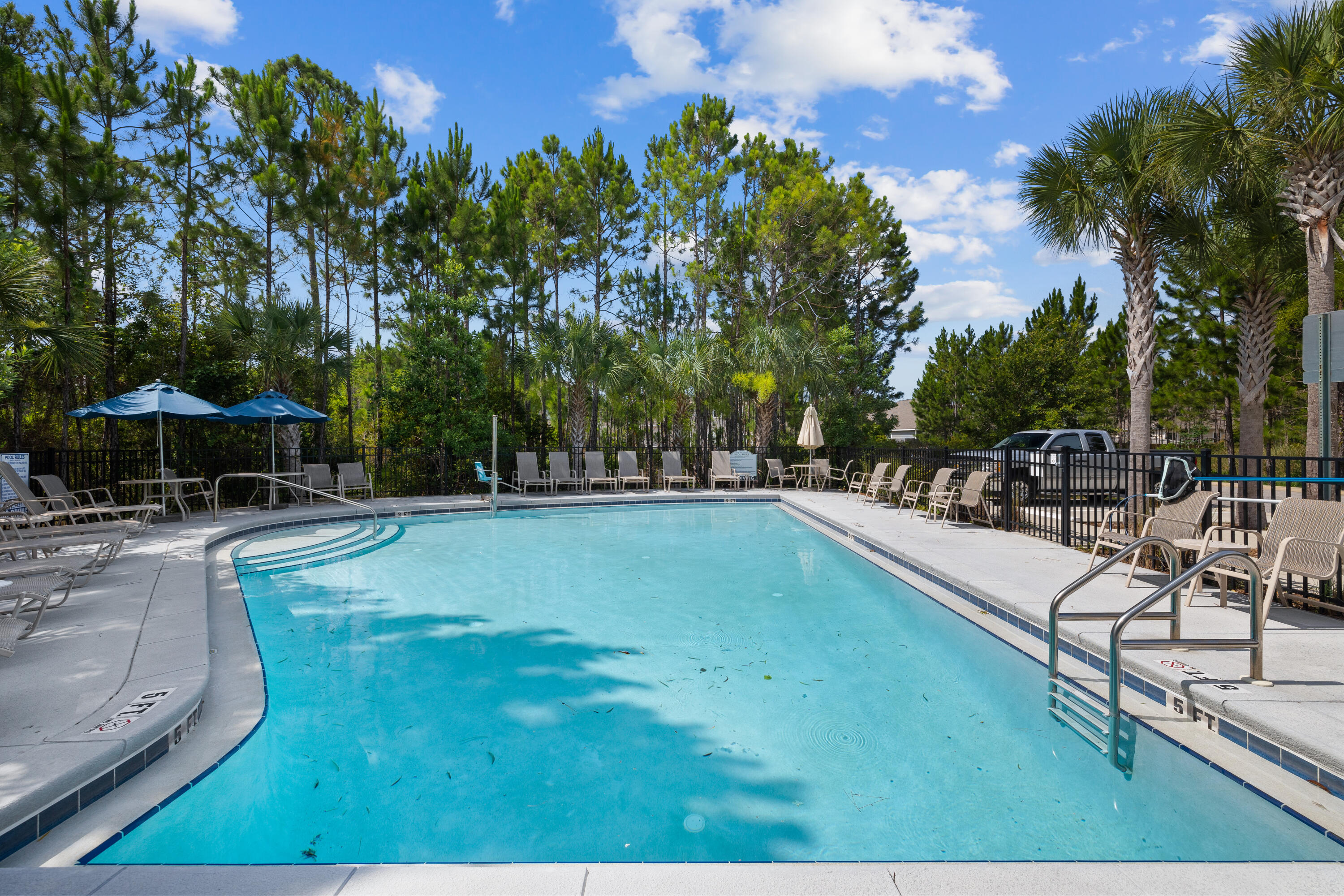Outdoor swimming pool with a curved shallow entry, lounge chairs, blue umbrellas, and surrounding pine trees.