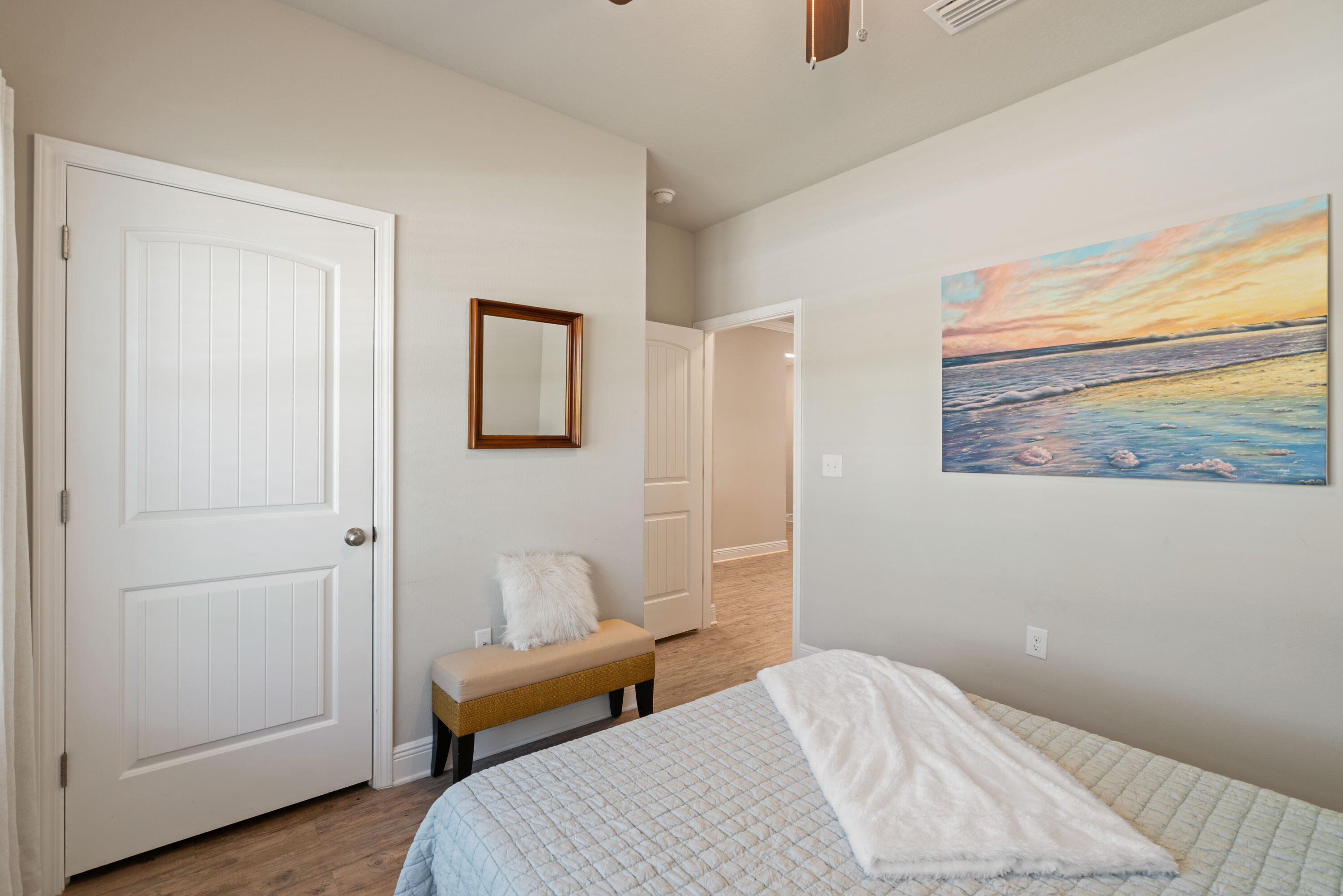 Bedroom with a quilt-covered bed, bench, white paneled door, and beach sunset painting.