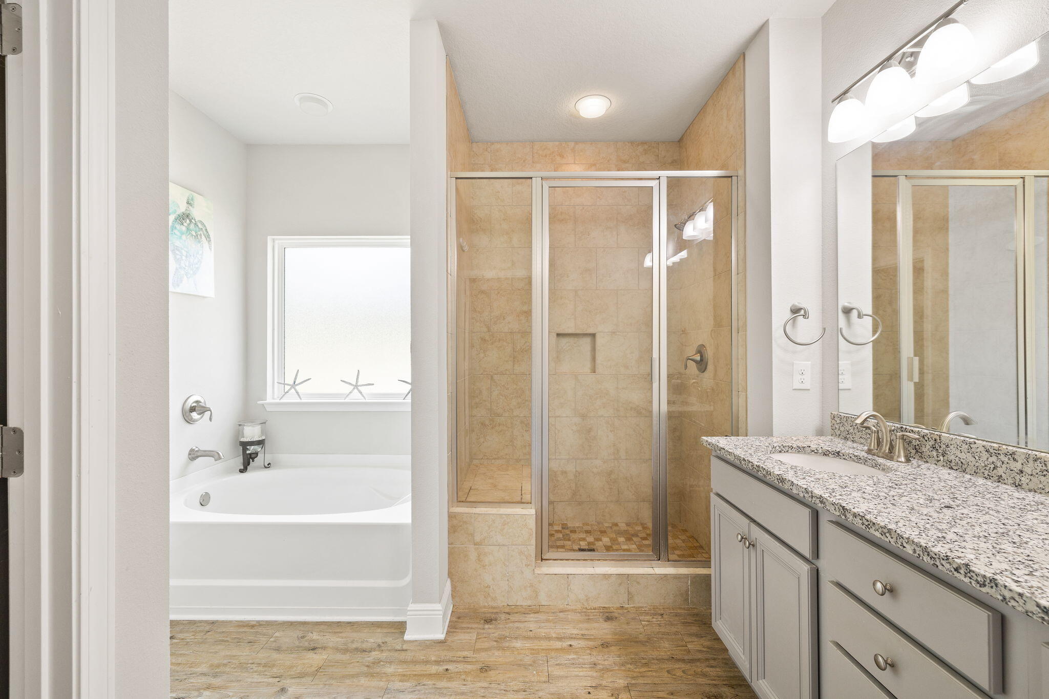 Bathroom with a white soaking tub, tan-tiled glass shower, and gray double vanity with granite countertop.