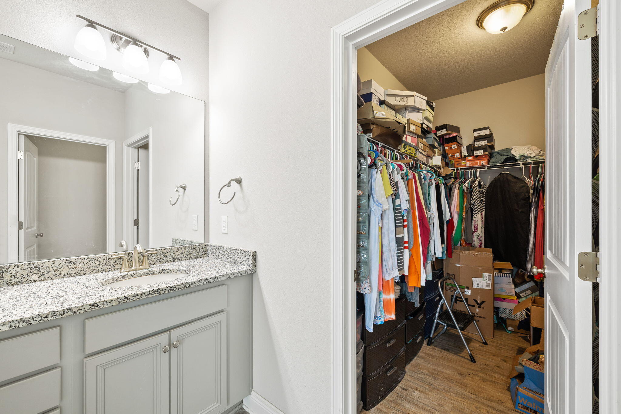 Bathroom vanity beside a crowded walk-in closet with hanging clothes and stacked boxes.