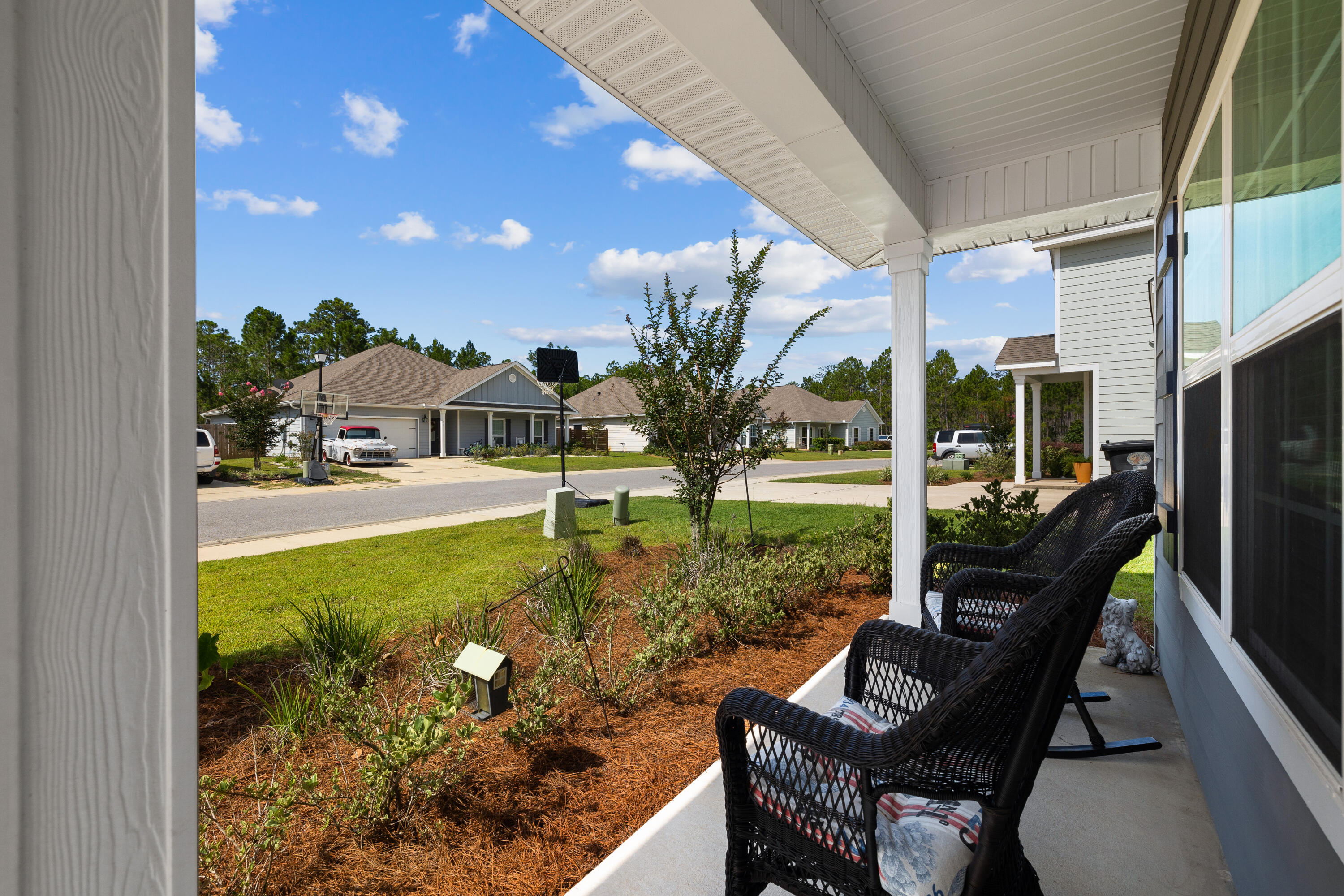 Covered front porch with black wicker rocking chairs overlooking a landscaped yard and neighboring homes.