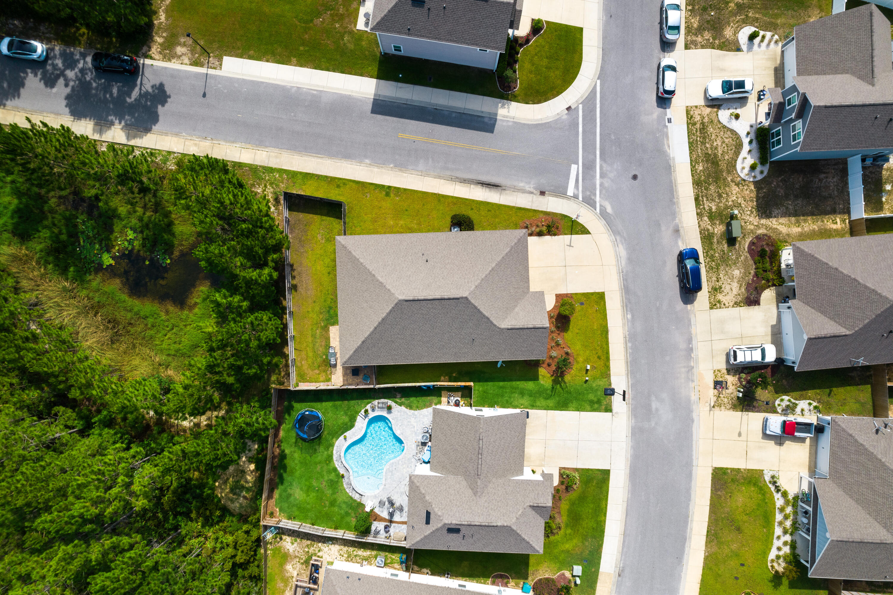 Aerial view of a corner-lot house with a fenced backyard and curving swimming pool beside dense trees.