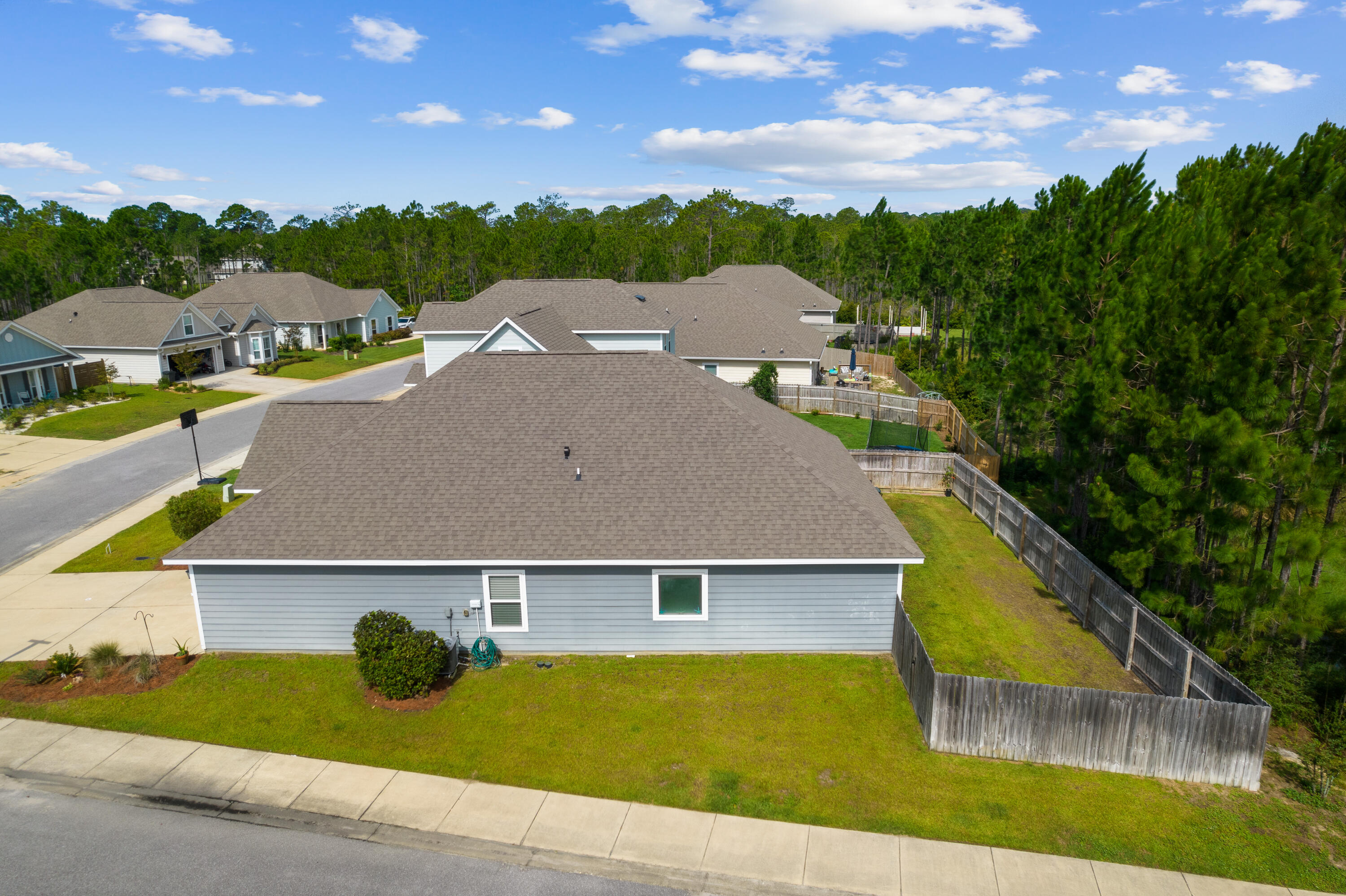 Aerial view of a gray-roofed home with a fenced lawn and wooded surroundings.