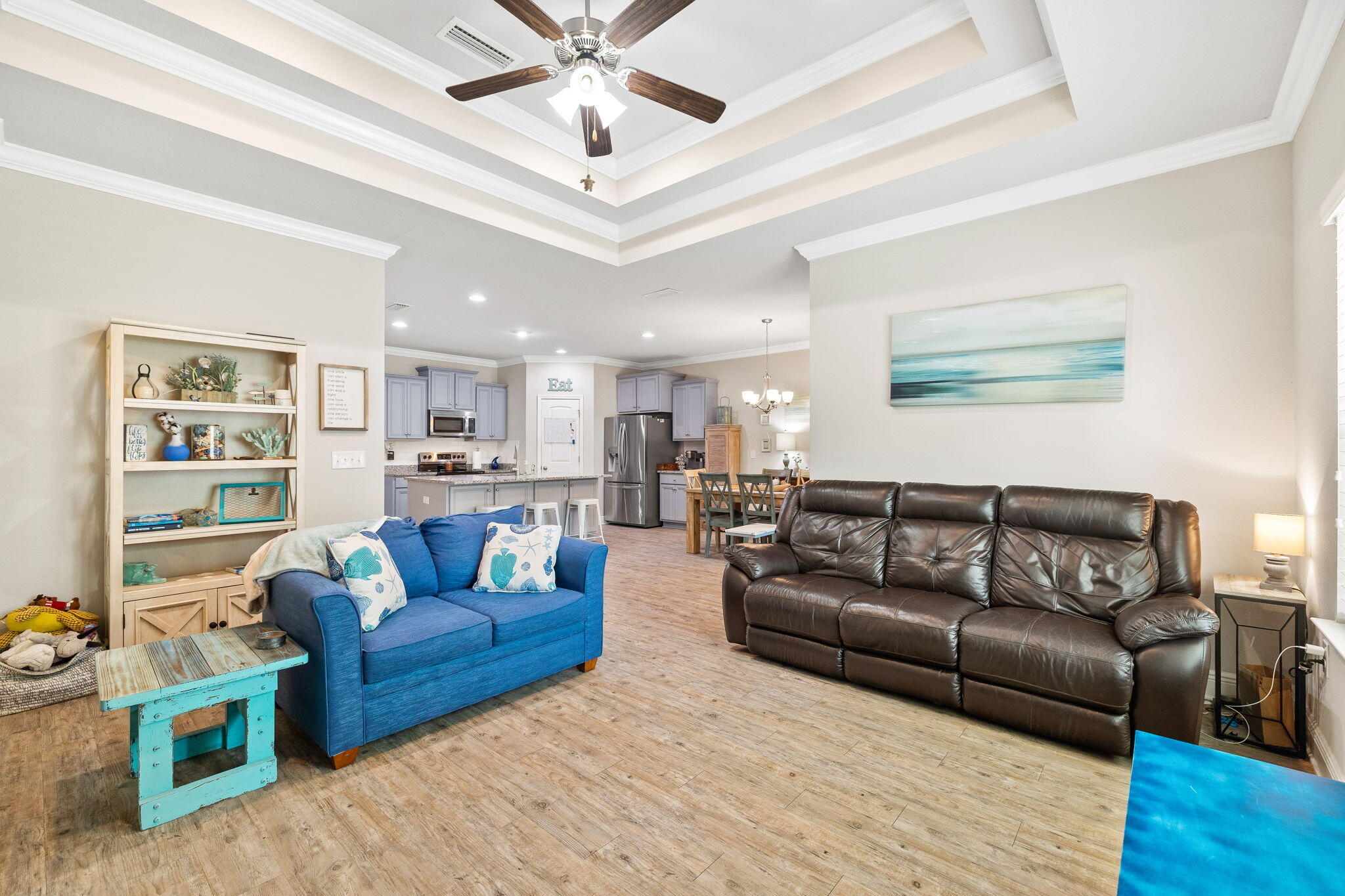 Open-plan living room and kitchen with a tray ceiling, blue loveseat, and dark leather sofa.