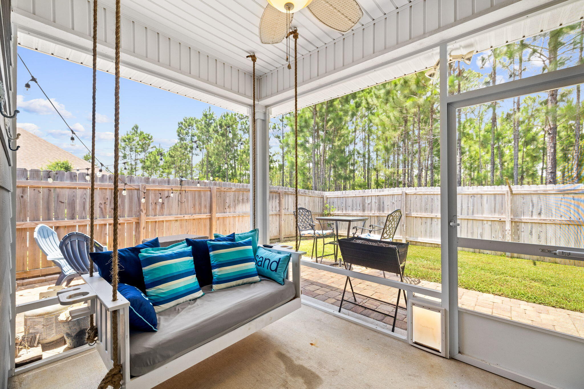 Screened porch with a rope-hung daybed overlooking a fenced yard and tall pine trees.