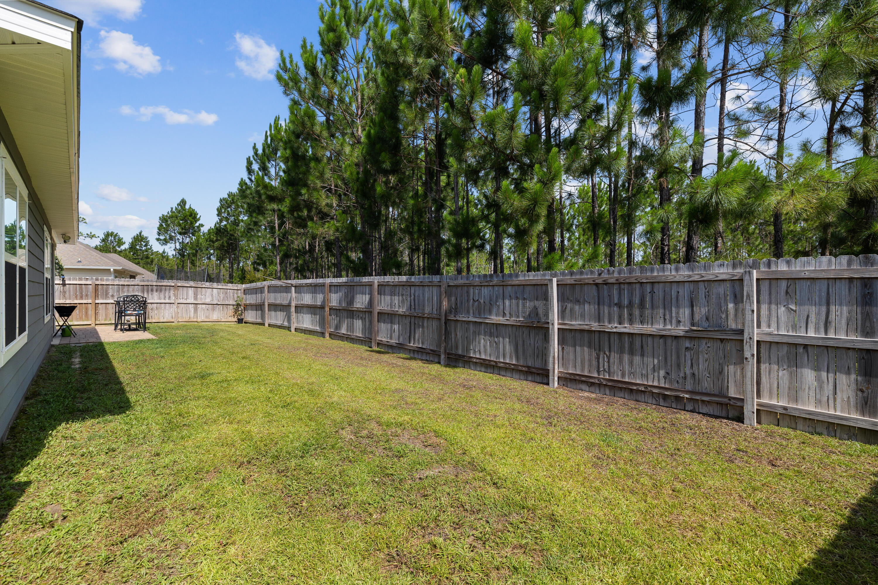 Fenced grassy backyard with a small patio and tall pine trees beyond the fence.