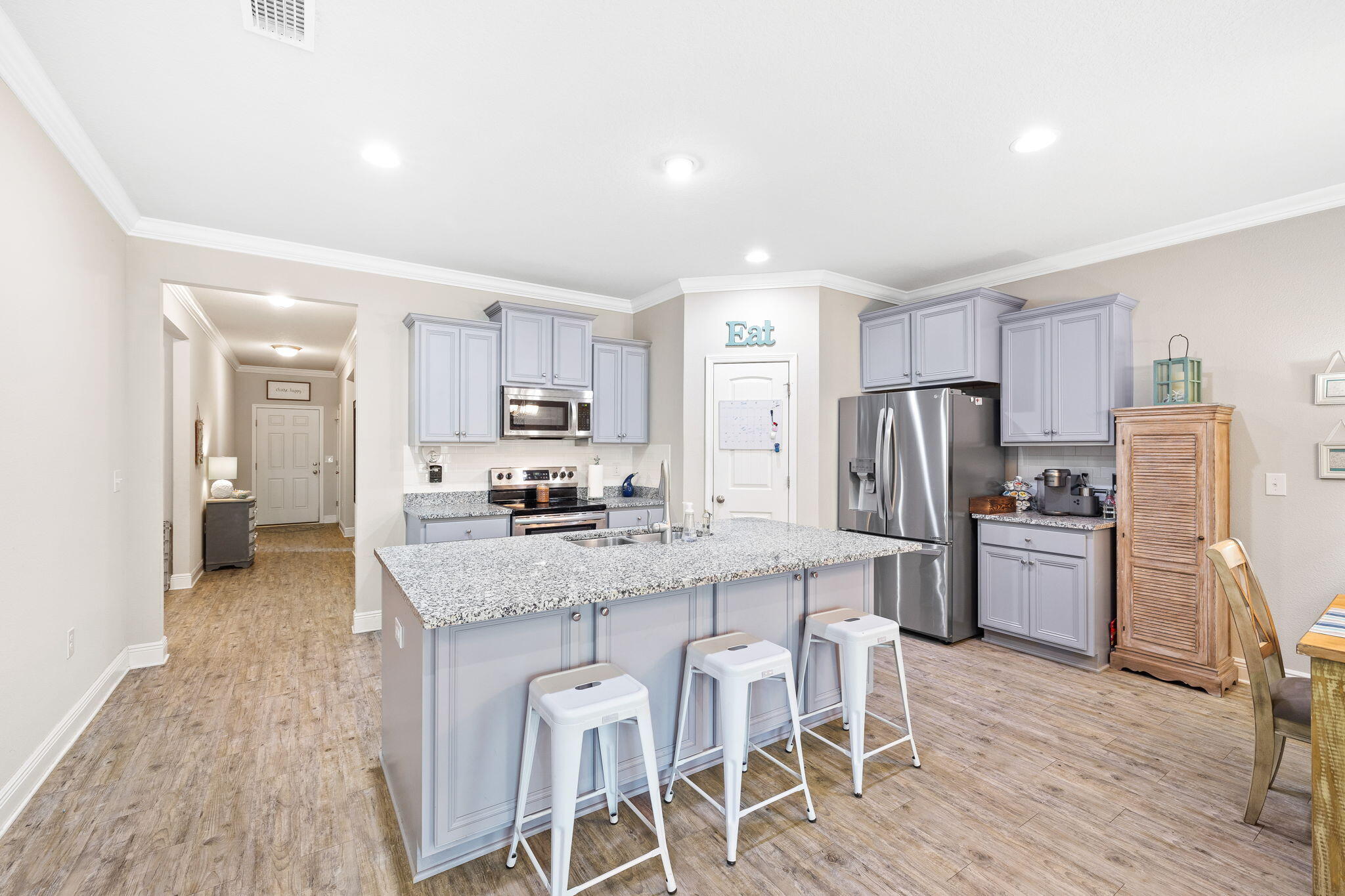 Kitchen with a large granite island, three white stools, gray cabinets, and stainless steel appliances.