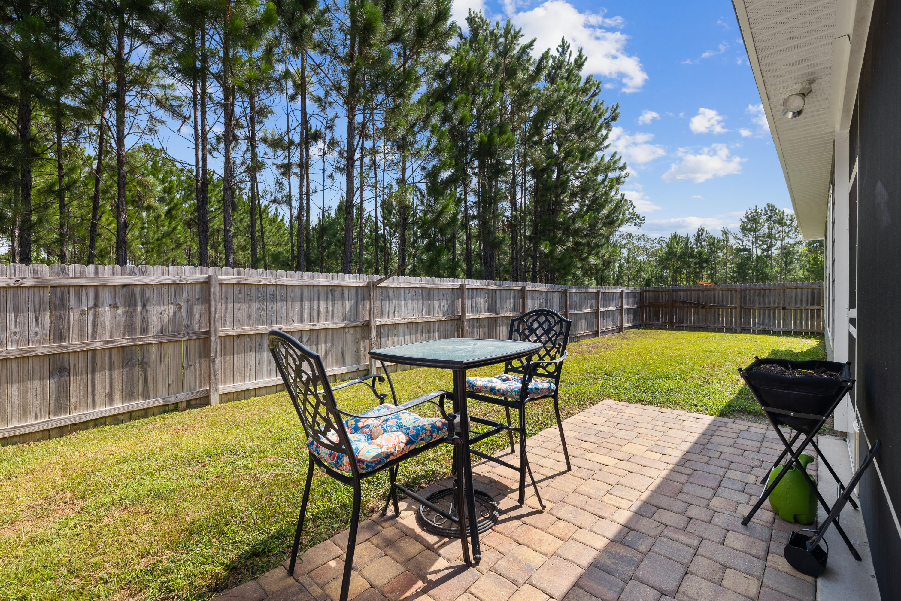 Backyard patio with brick pavers, metal seating, a fenced lawn, and pine trees.
