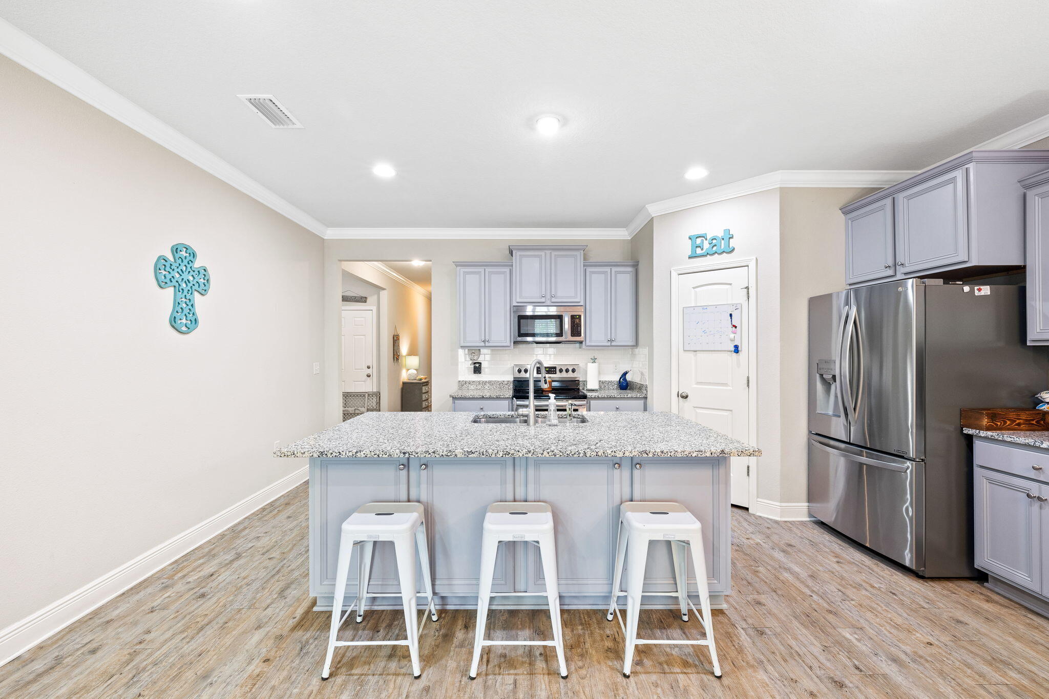 Kitchen with a large granite island, three white stools, pale blue-gray cabinets, and stainless steel appliances.
