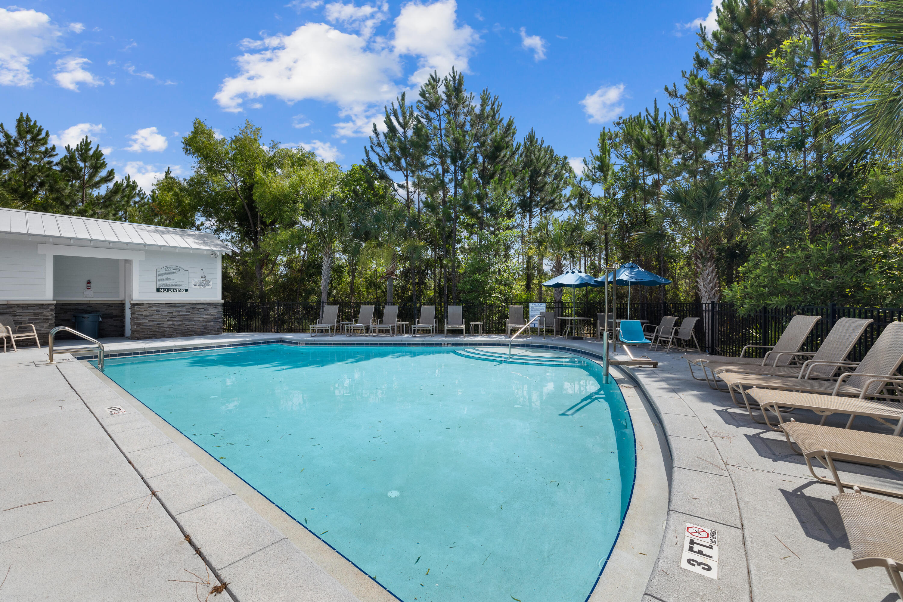 Community swimming pool with turquoise water, lounge chairs, blue umbrellas, and a tree-lined fence.