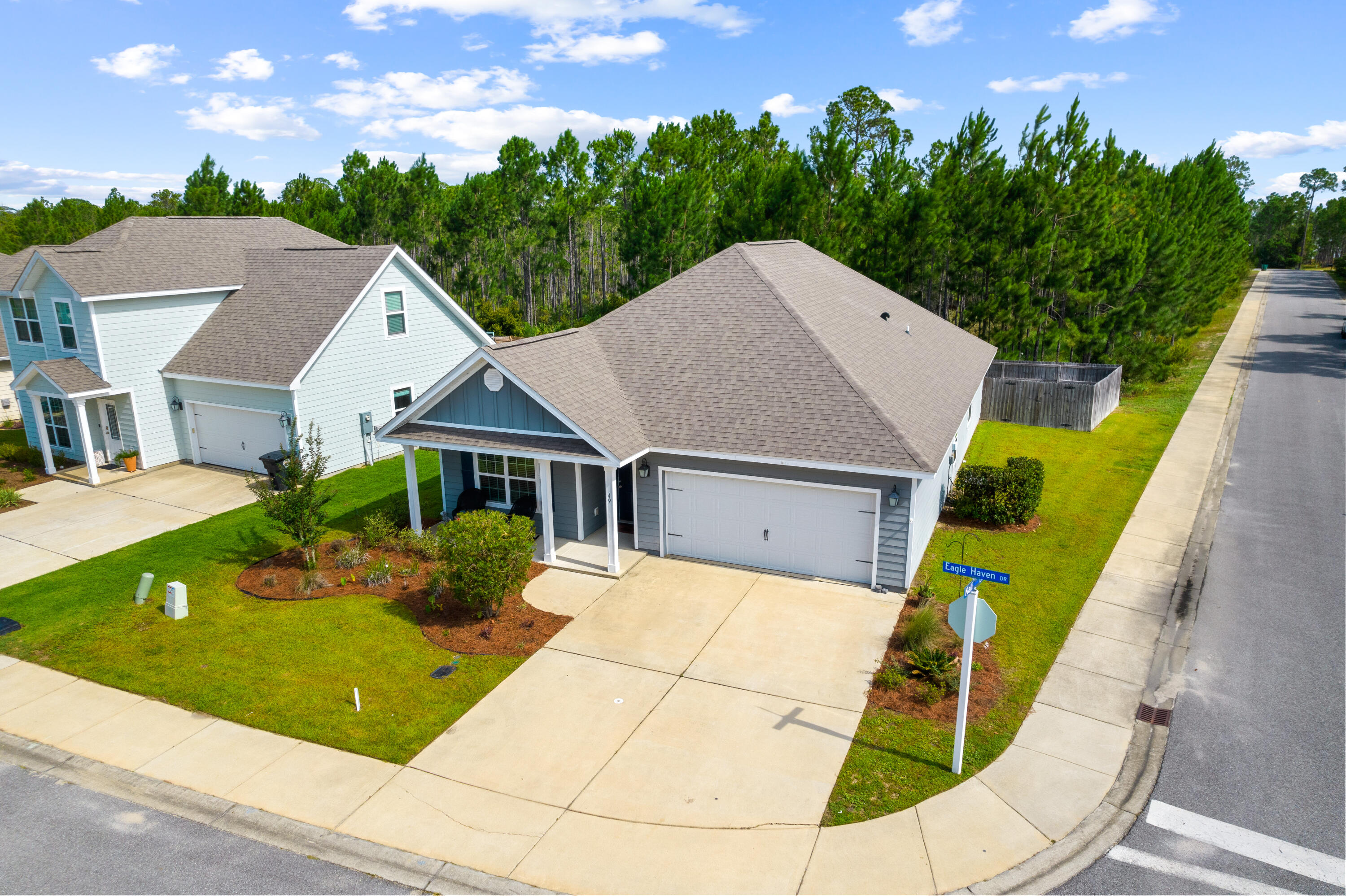 Aerial view of a blue-gray corner-lot home with a covered porch, two-car garage, and wooded backdrop.