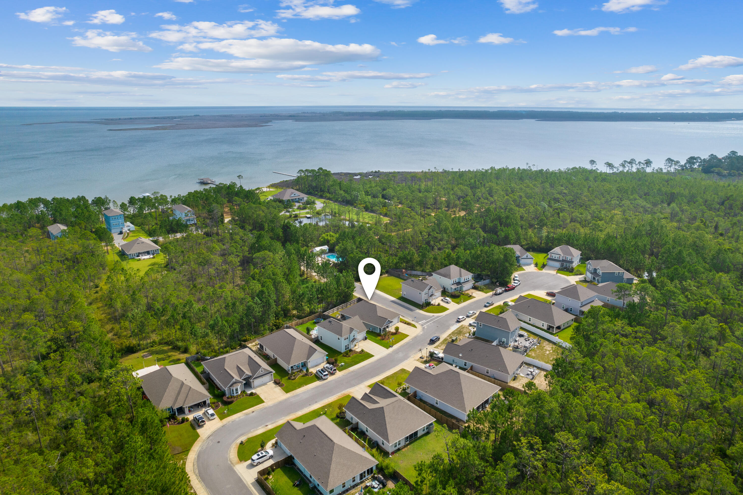 Aerial view of homes beside dense pine woods and a broad bay, with a map pin marking one home.