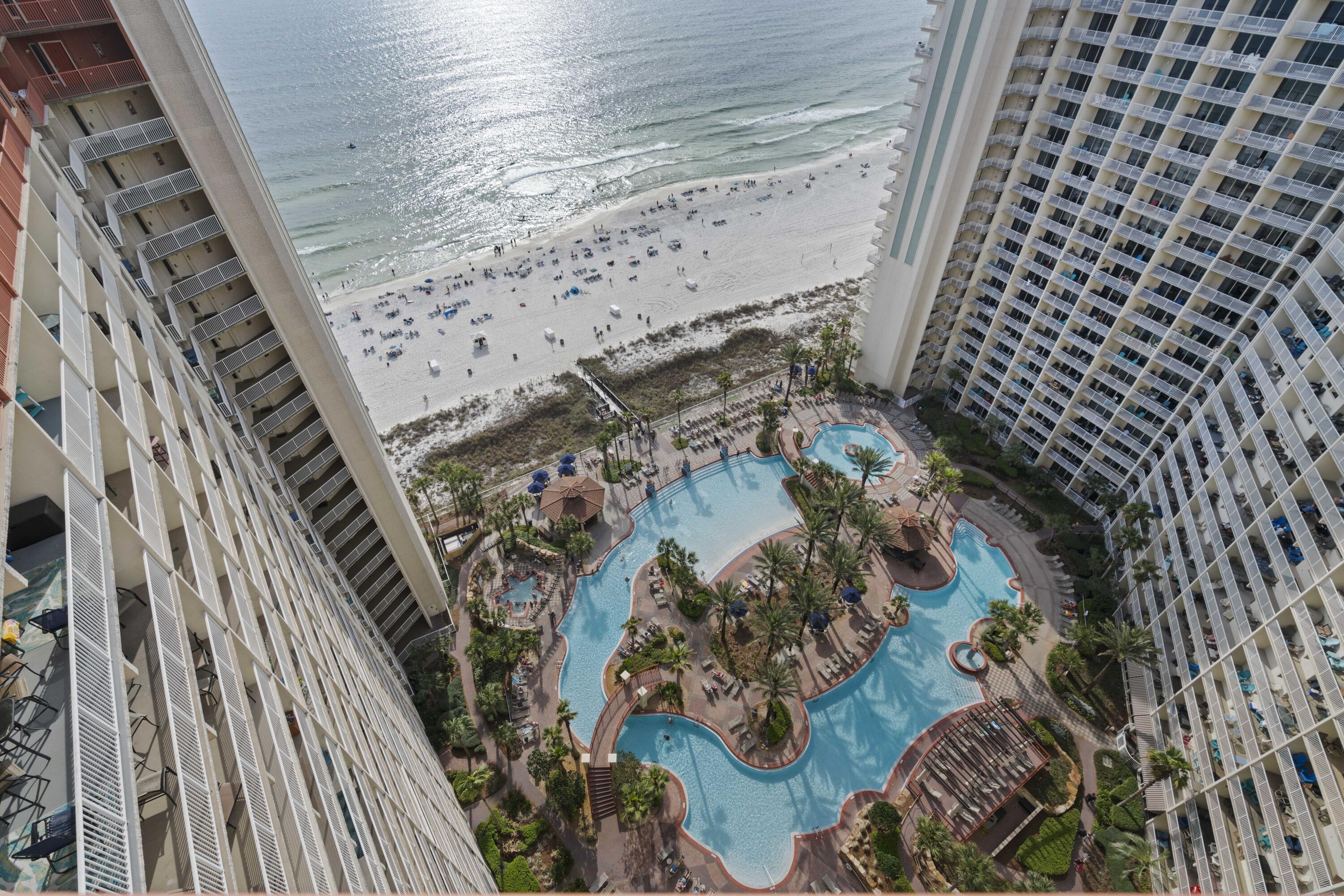 Aerial view of a winding resort pool between beachfront towers, with the beach and ocean beyond.