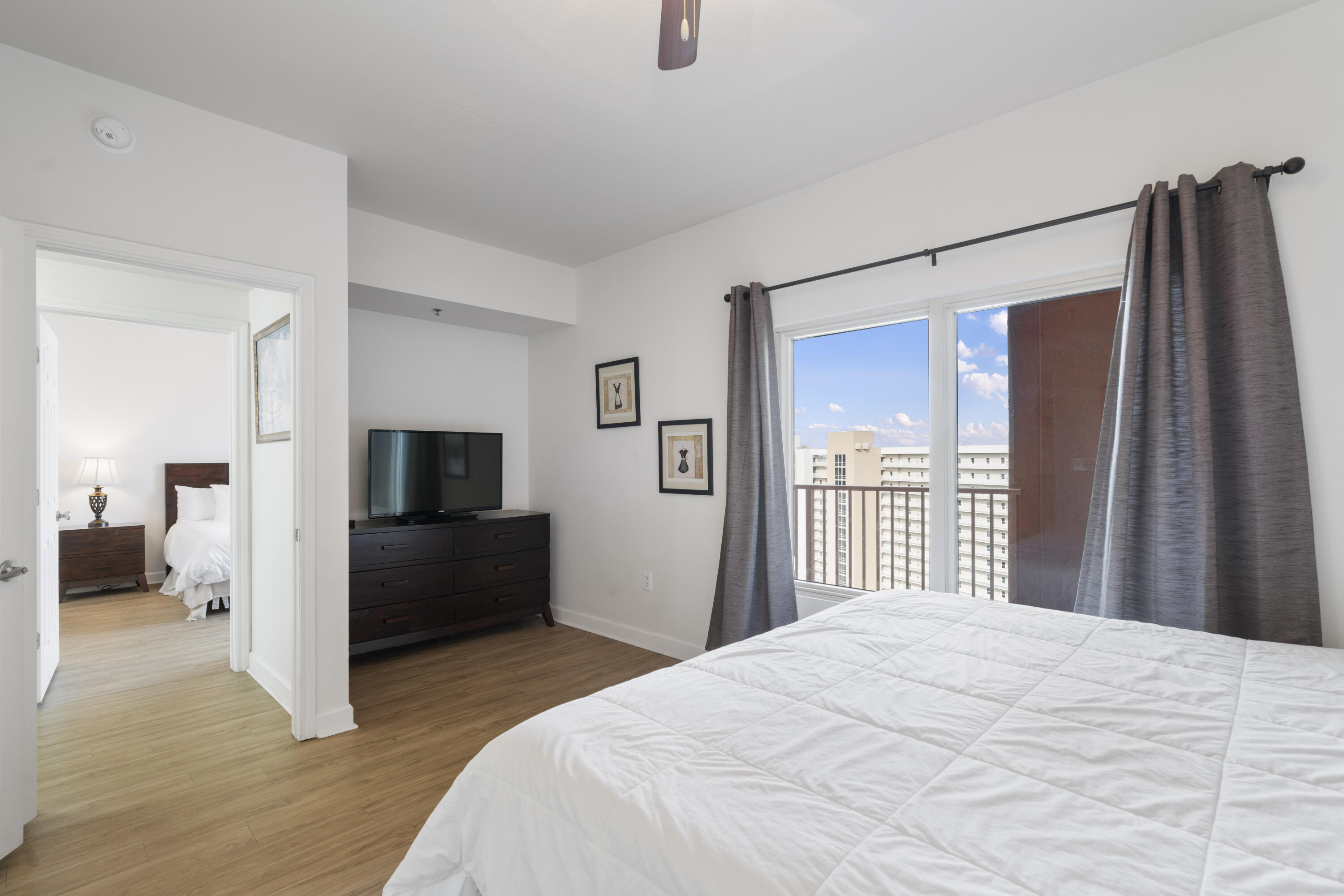 Bedroom with a white bed, dark dresser and TV, and a window overlooking nearby high-rise buildings.