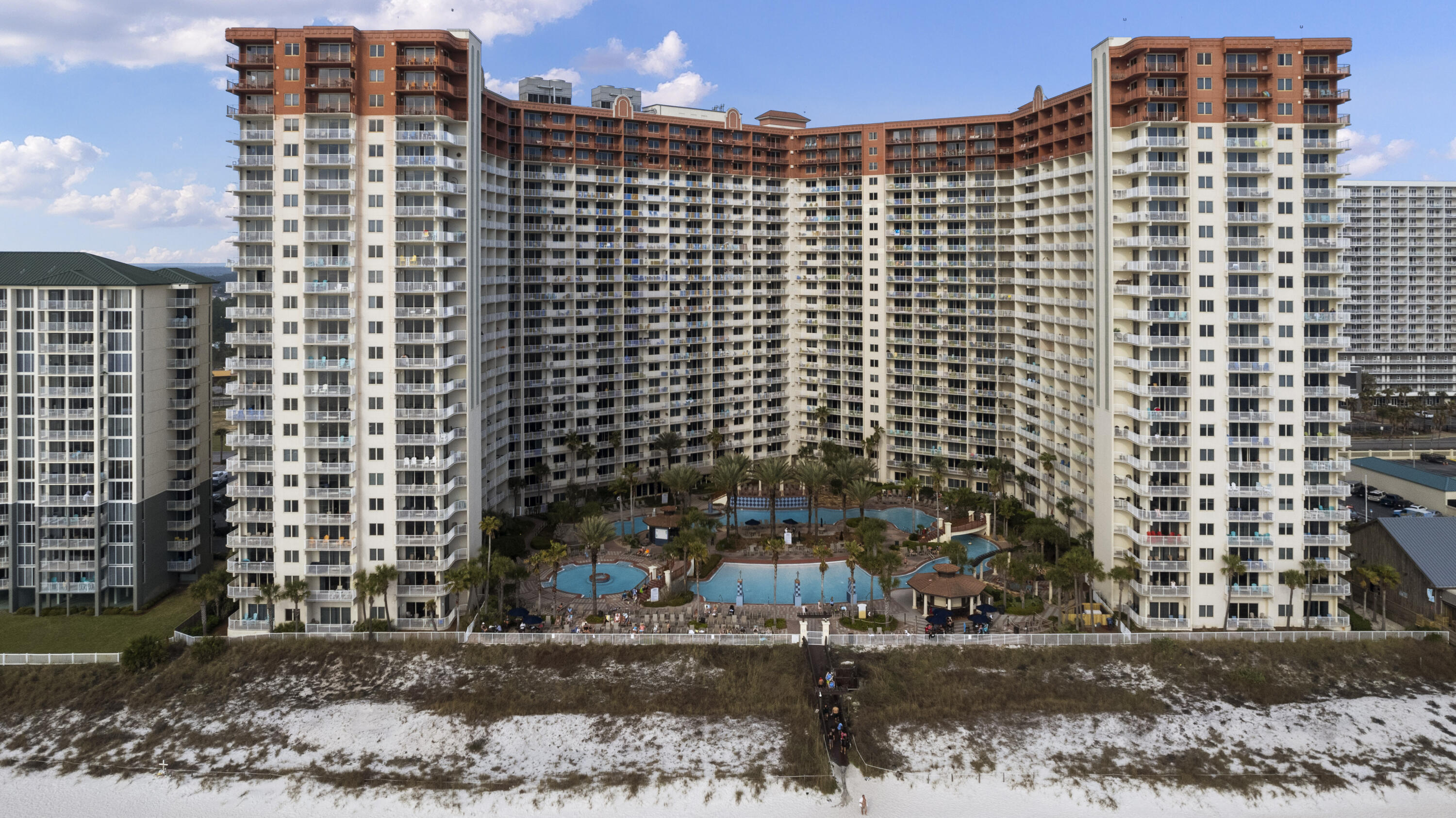 Aerial view of beachfront condominium towers surrounding multiple pools, with a sandy beach in front.