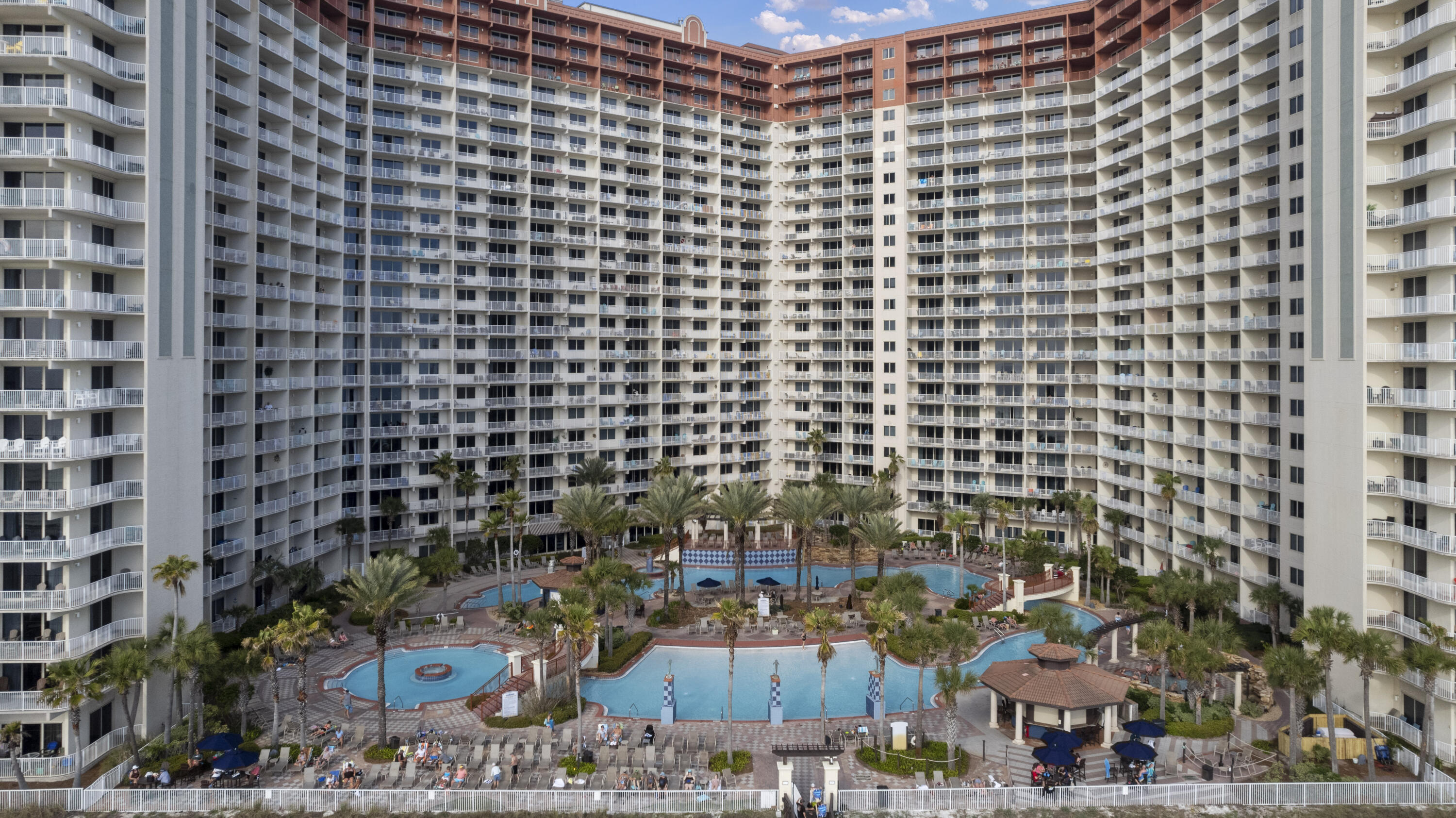 Condominium courtyard with multiple swimming pools, palm trees, and surrounding multistory buildings.