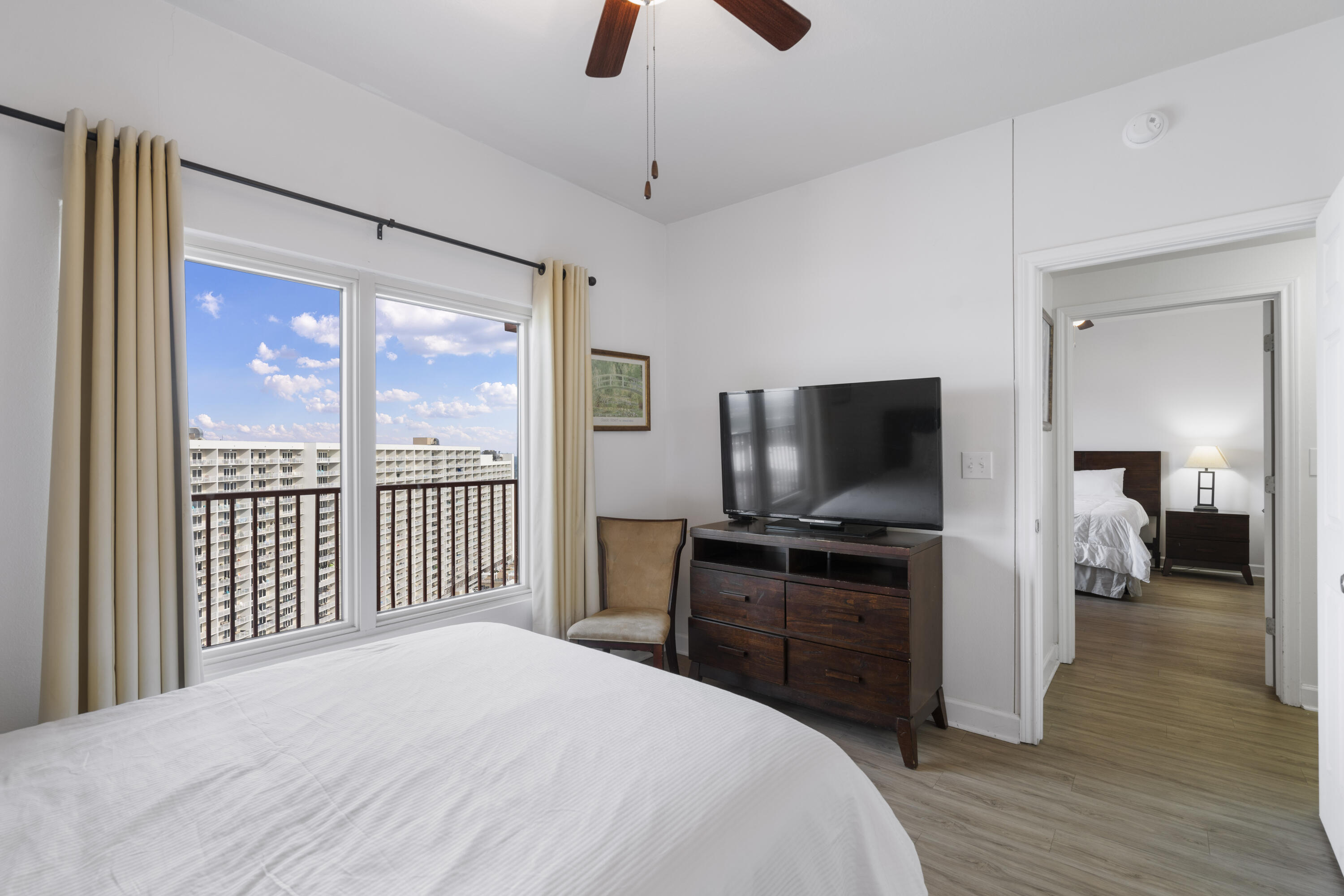 Bedroom with a white bed, dark wood dresser and TV, and windows overlooking neighboring high-rises.