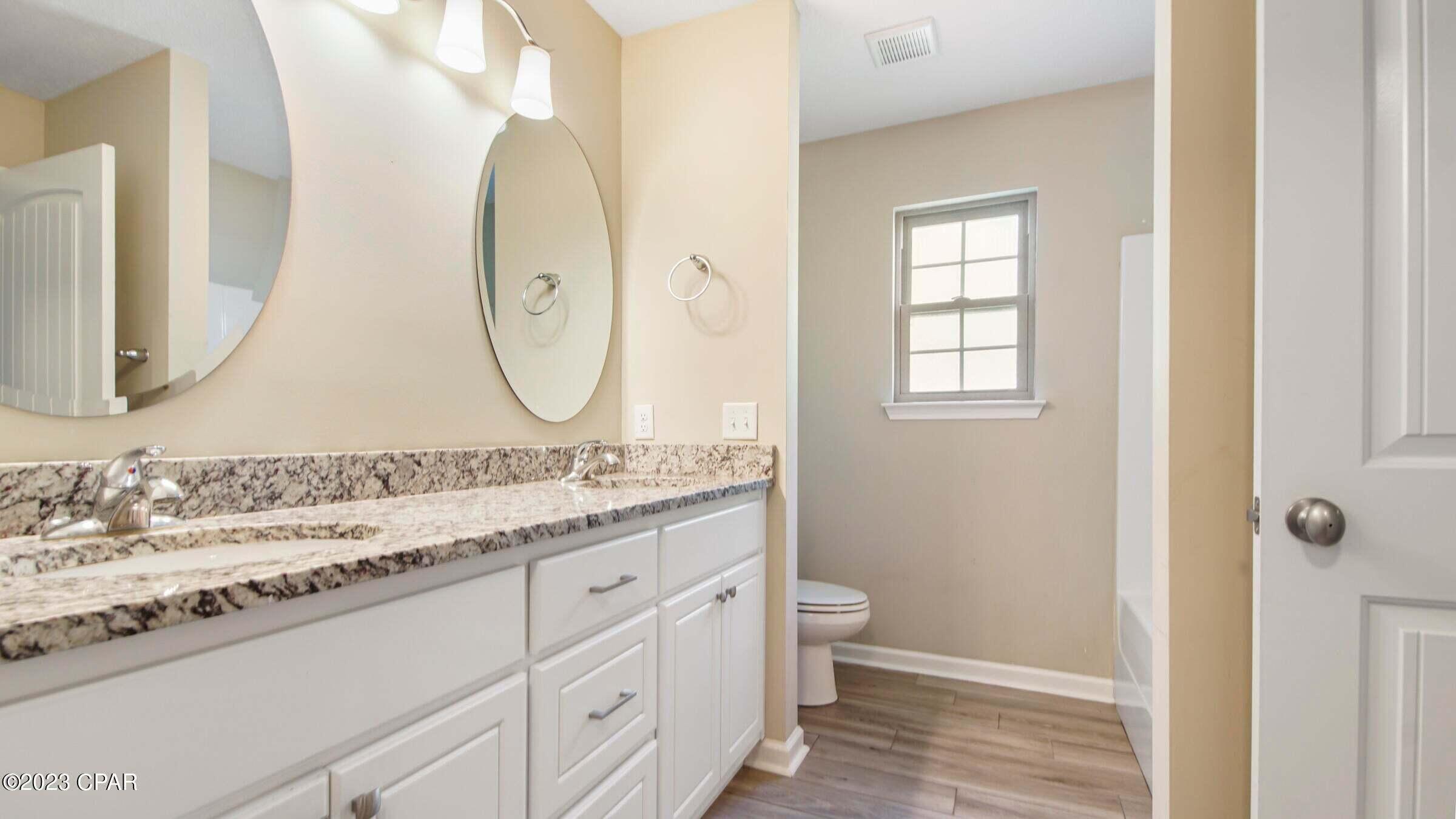 Bathroom with a granite double vanity and oval mirrors, viewed toward a toilet beneath a window.