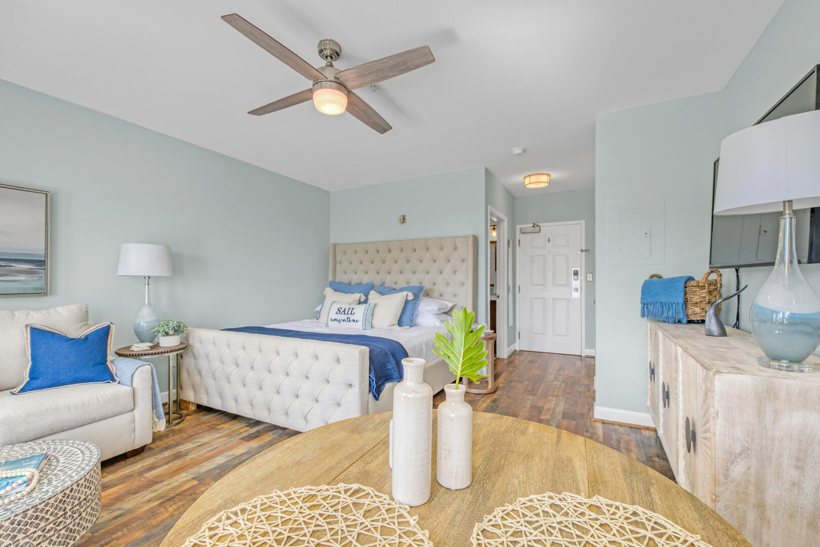Staged bedroom with a tufted white bed, blue accents, cream seating, and wood-look flooring.